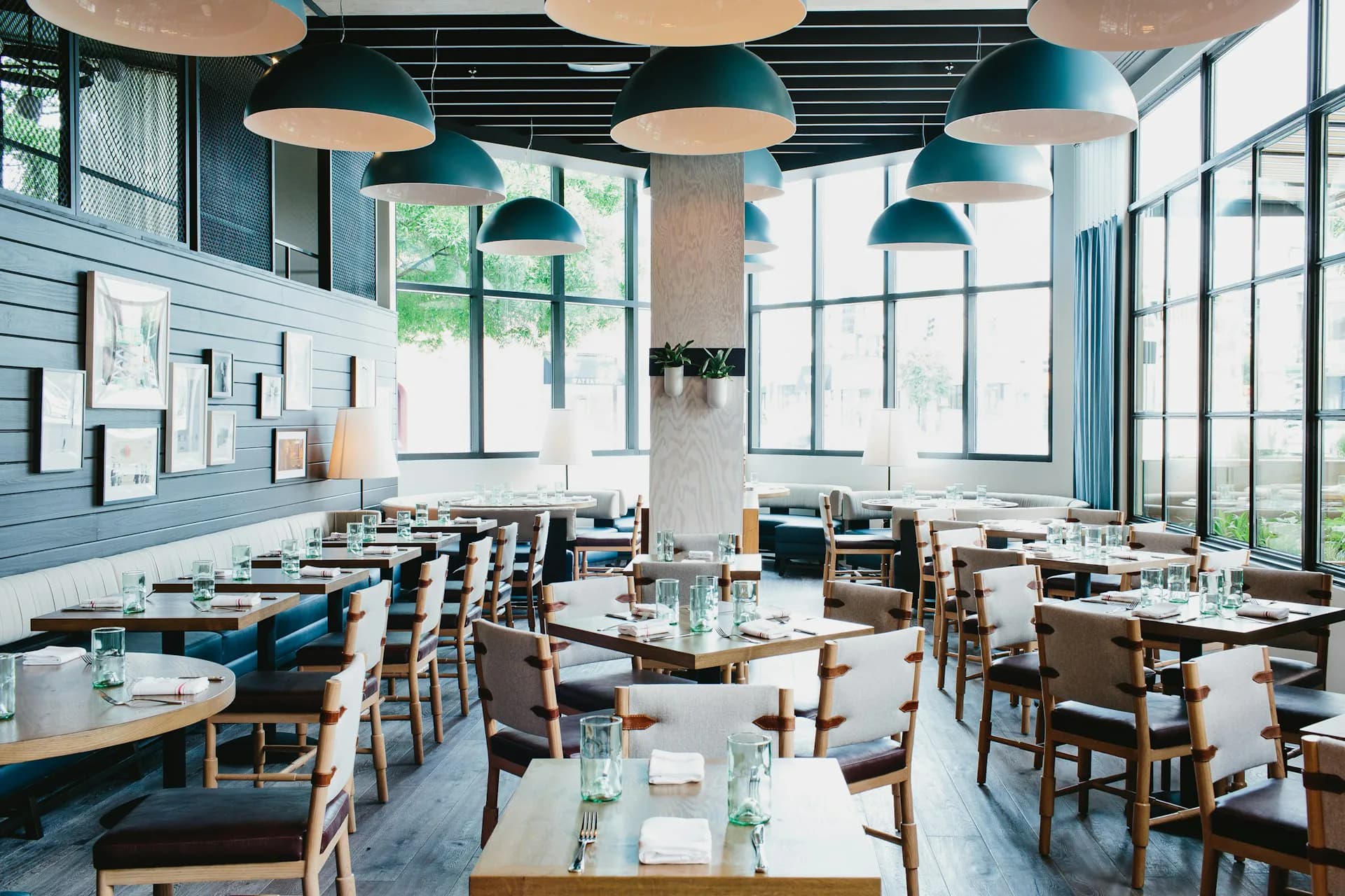 Contemporary restaurant interior with blue-green walls, wooden chairs, round tables, pendant lighting, and floor-to-ceiling windows by Meyer Davis Studio Inc.