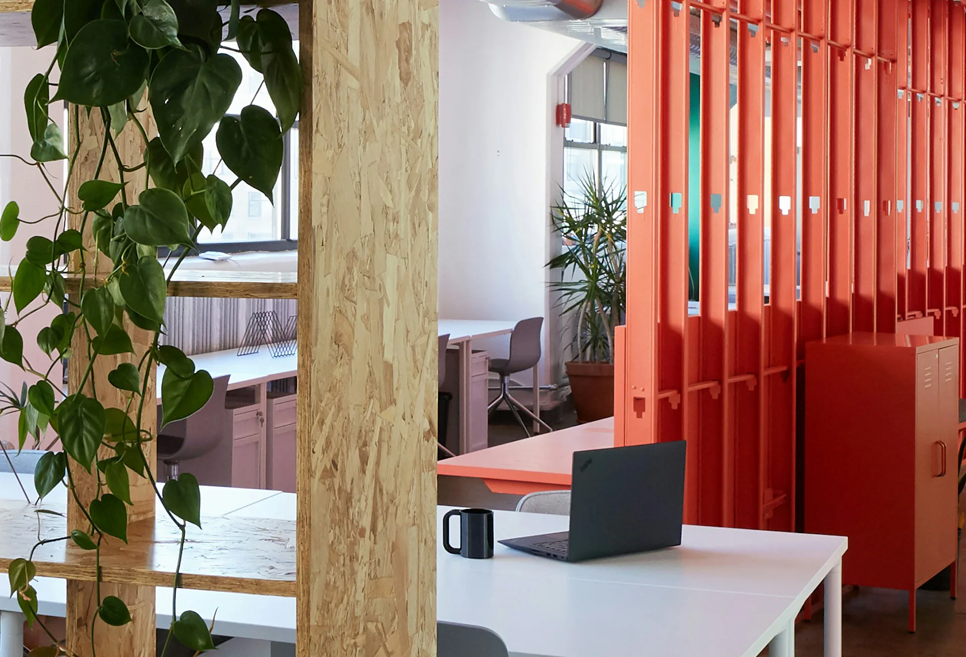 Modern office interior with wooden column, white desk, black laptop, vibrant red vertical louvers, and green plant accents by MGDK Architects.