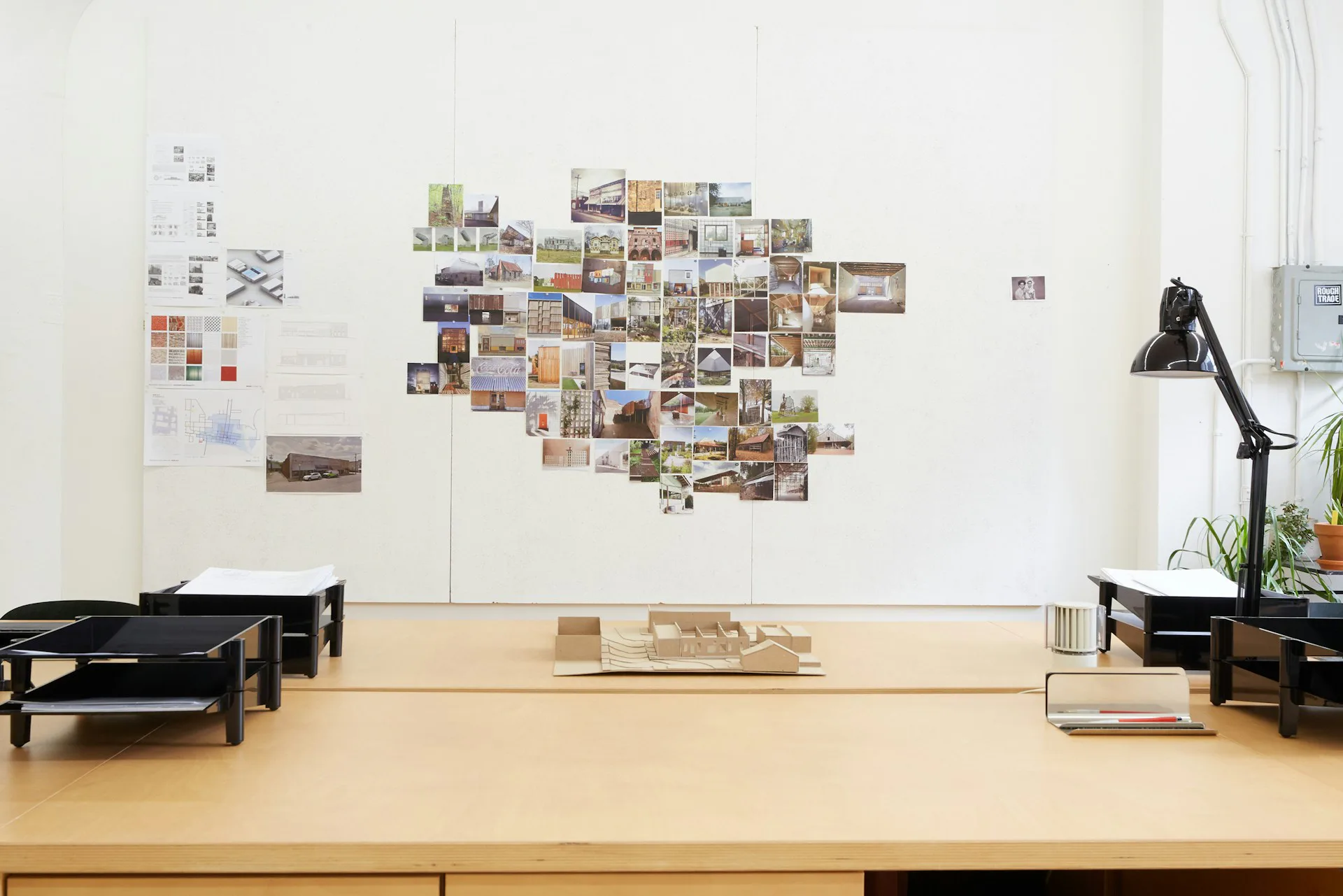 Design studio workspace with wooden desk, black task chairs, wall-mounted mood board with photographs and sketches, and desk accessories by MGDK Architects.