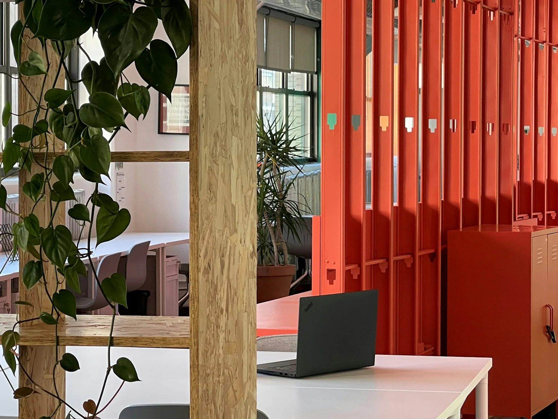 Modern office interior with red corrugated metal storage, natural wood column, green climbing plants, pink flooring, and minimalist desk setup by MGDK Architects.