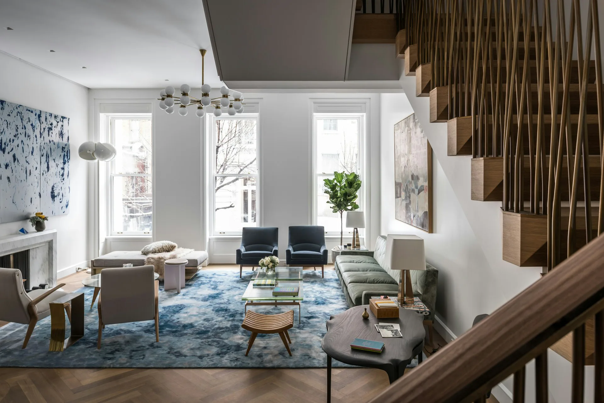 Bright, multi-level living space with blue area rug, mid-century modern seating, large windows, and wooden staircase details by Michael K Chen Architecture.