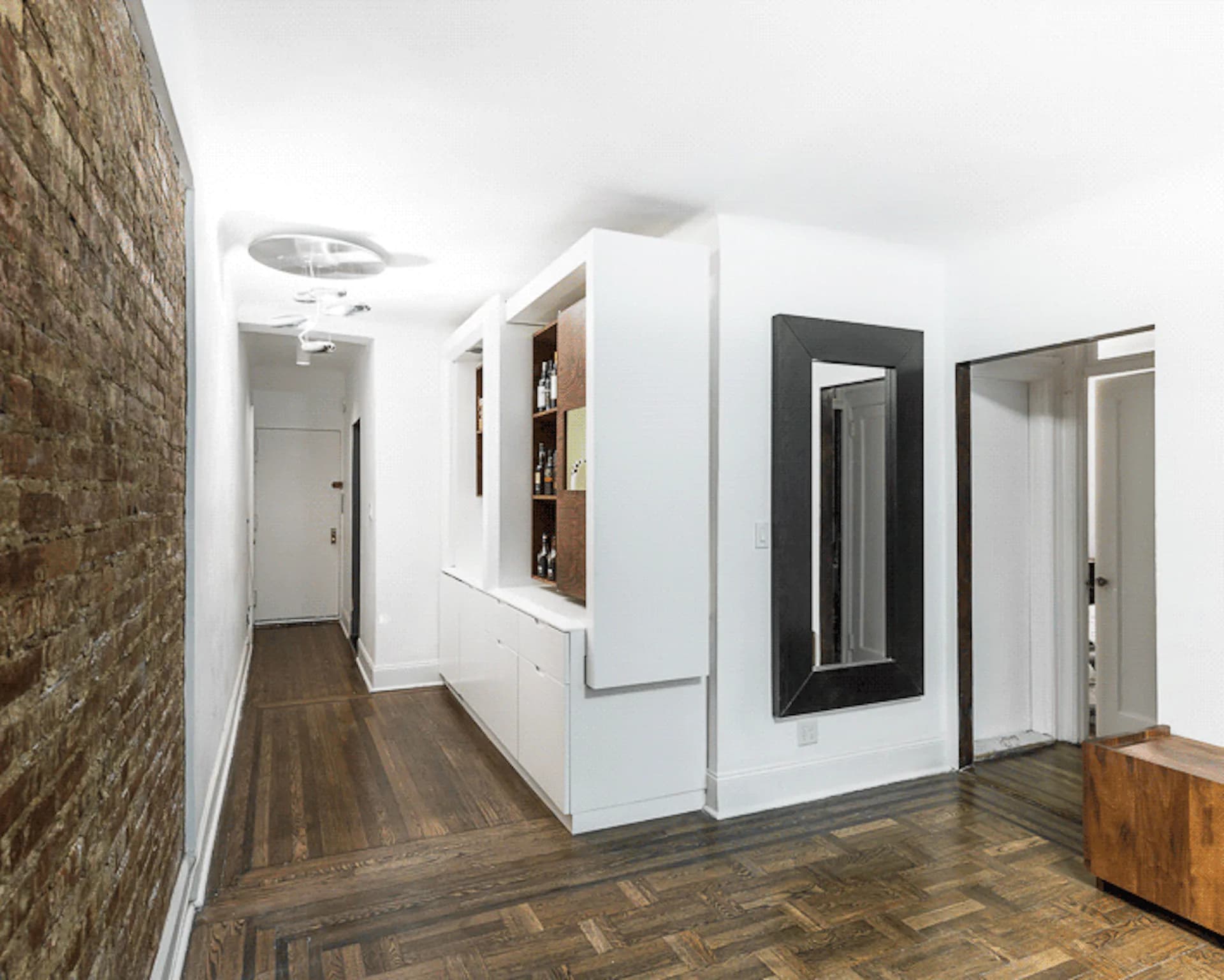 Modern apartment hallway with white built-in cabinetry, dark brick accent wall, herringbone wood flooring, and minimalist black-framed mirrors by Michael K Chen Architecture.