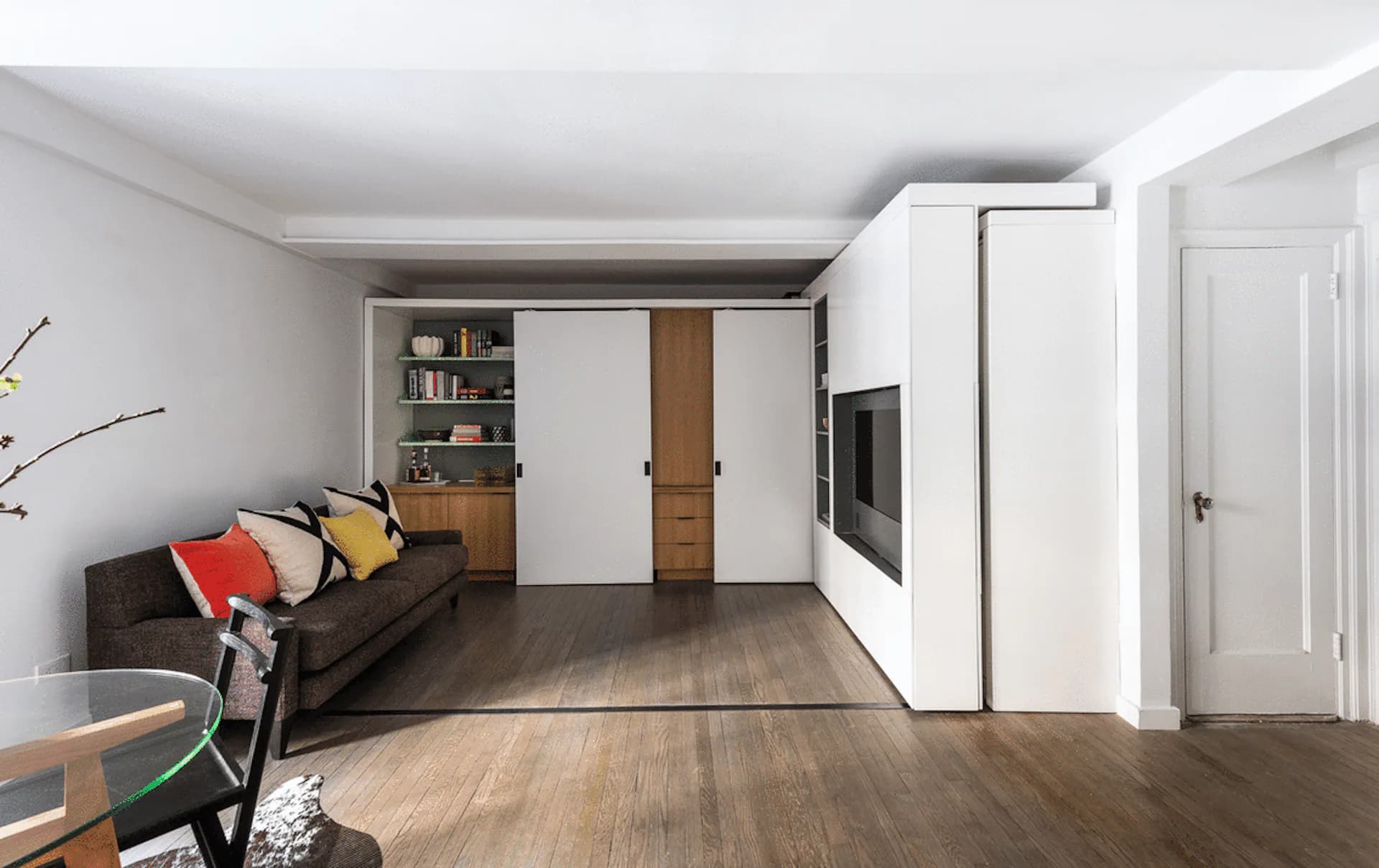 Modern living room rendering with white sliding wardrobe, wooden shelving, dark sofa, and wood flooring by Michael K Chen Architecture.