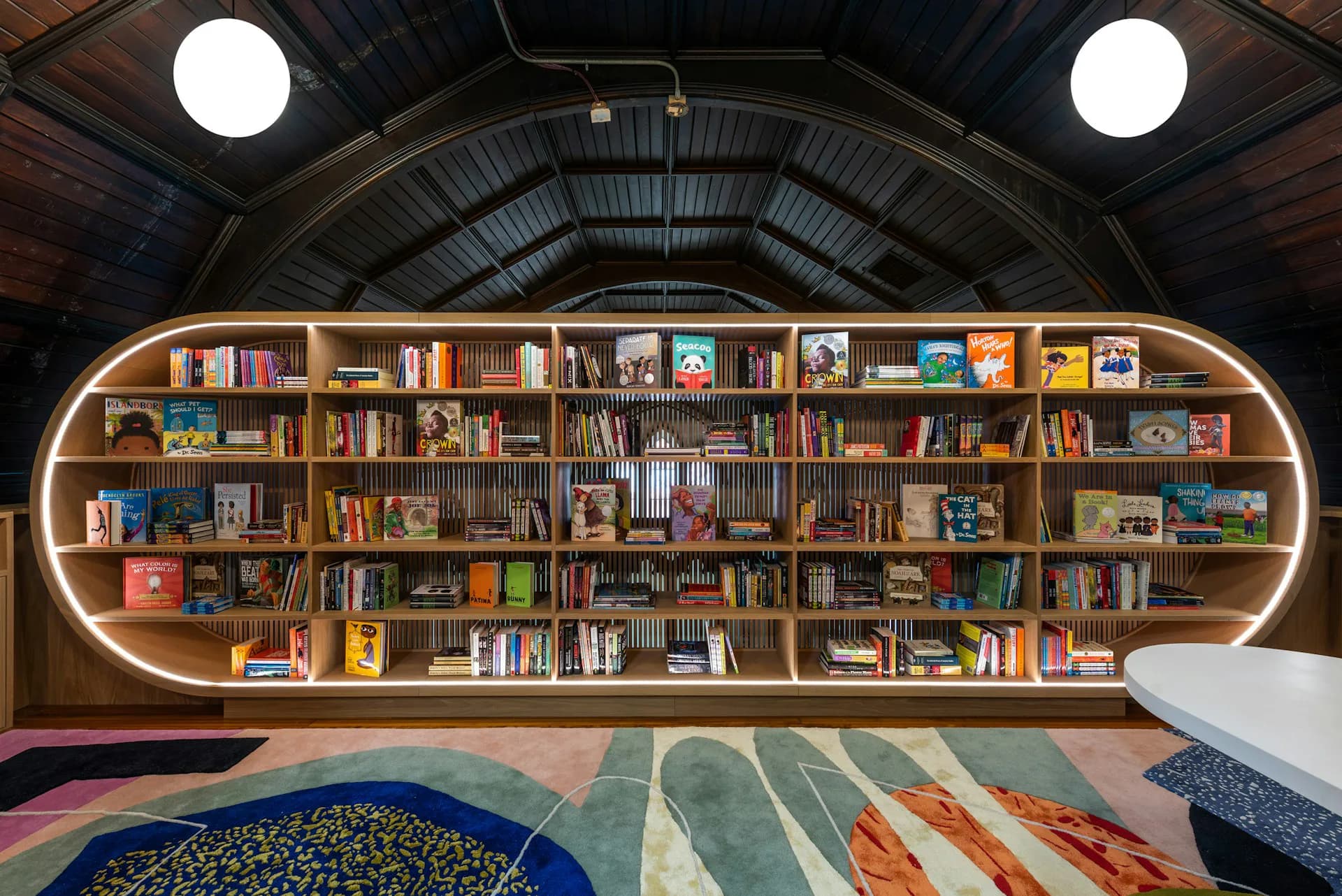 Modern library interior featuring a curved built-in bookshelf wall with warm wood tones and integrated lighting, colorful patterned rugs, and dramatic arched ceiling by Michael K Chen Architecture.