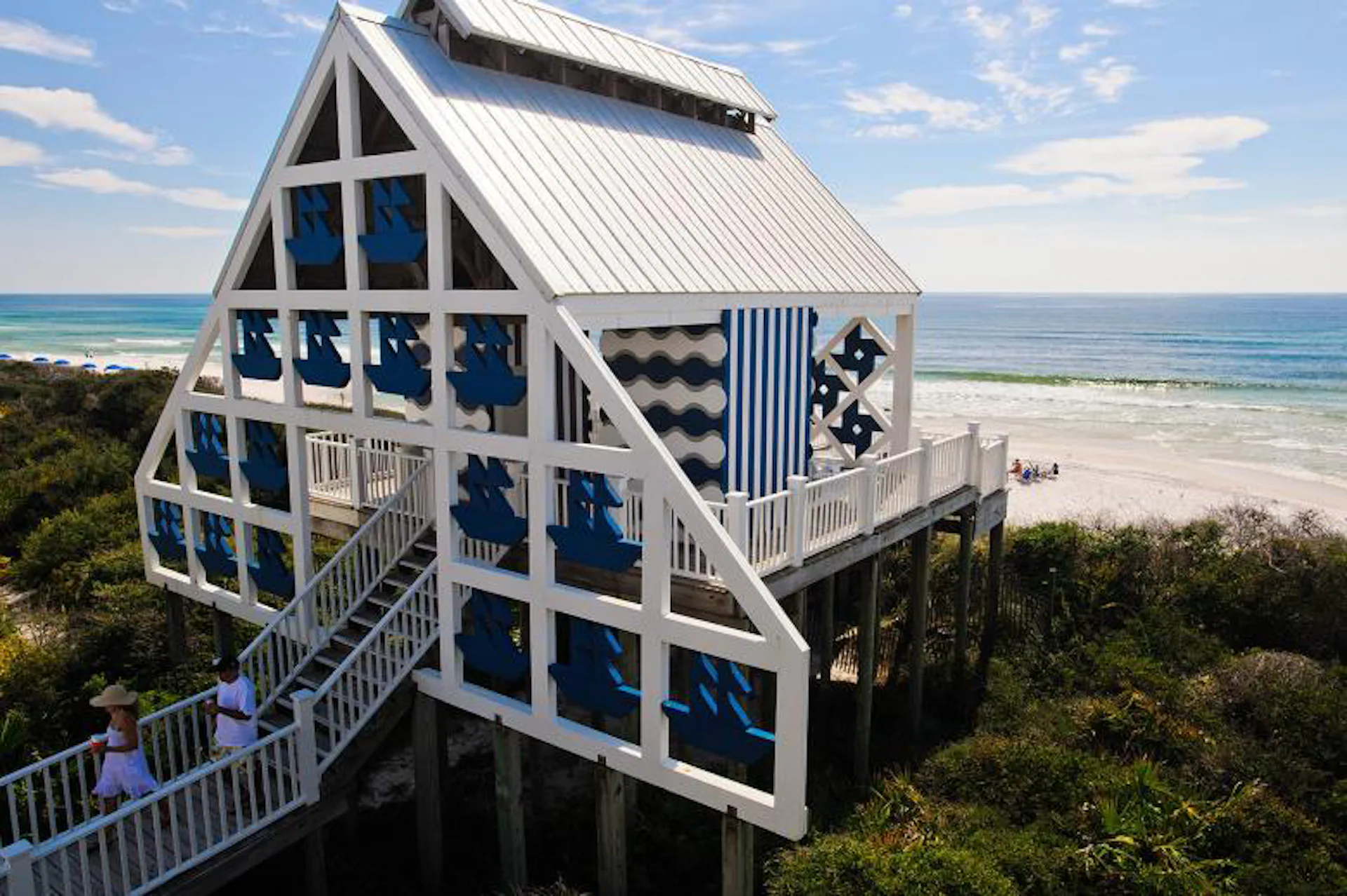 Beachfront A-frame house with white and blue exterior, elevated on stilts above coastal vegetation, featuring geometric window patterns and metal railings by Michael McDonough Architect.