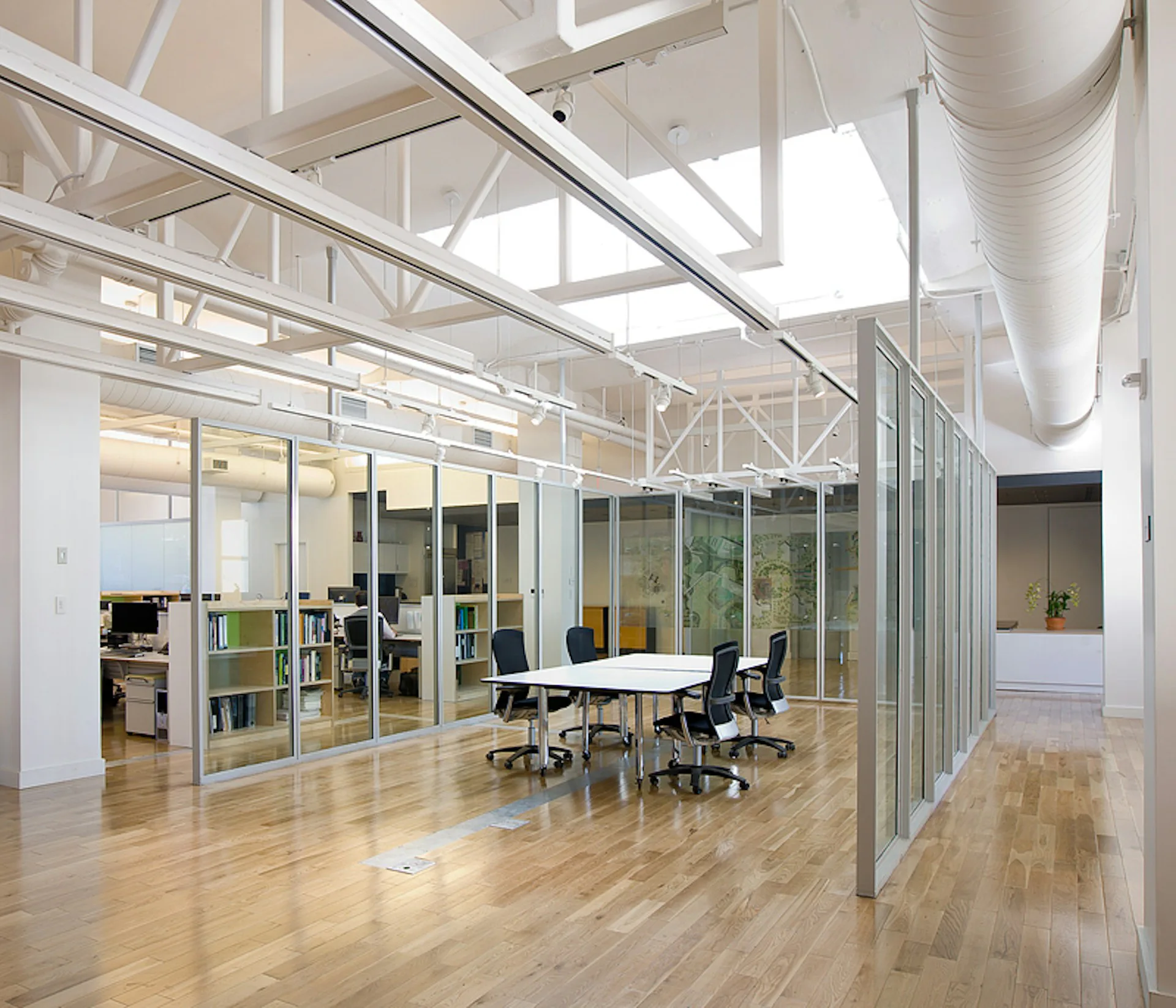 Bright open-plan office with glass-partitioned meeting room, wooden flooring, skylights, and exposed ceiling systems by Michielli + Wyetzner Architects.
