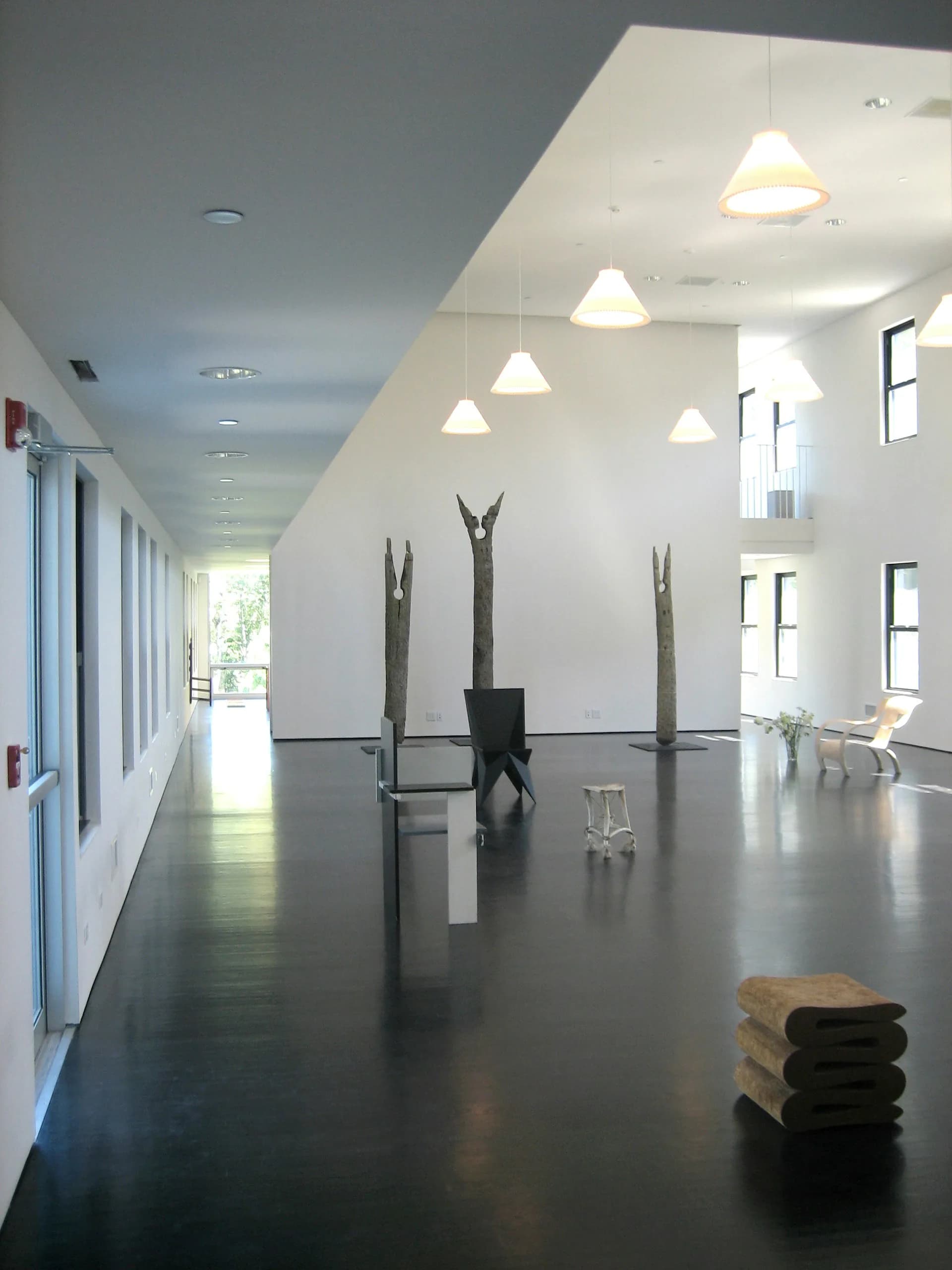 A bright, minimalist gallery space with soaring white walls, pendant lighting, dark flooring, and sculptural metal pieces displayed, designed by Michielli + Wyetzner Architects.