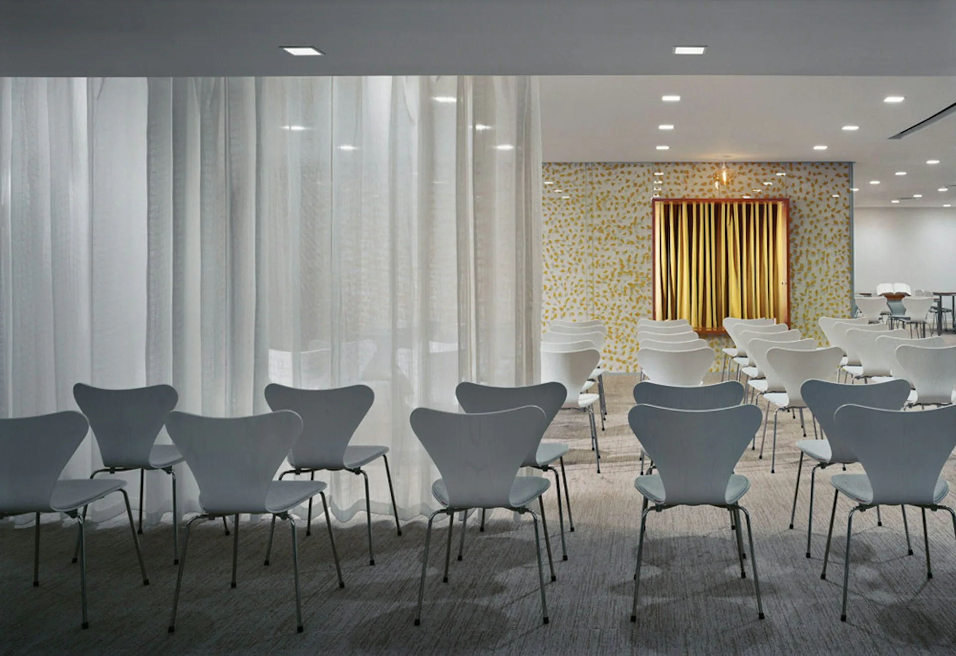 Modern conference space with gray wingback chairs arranged in rows, golden curtains, textured accent wall, and recessed lighting by Michielli + Wyetzner Architects.