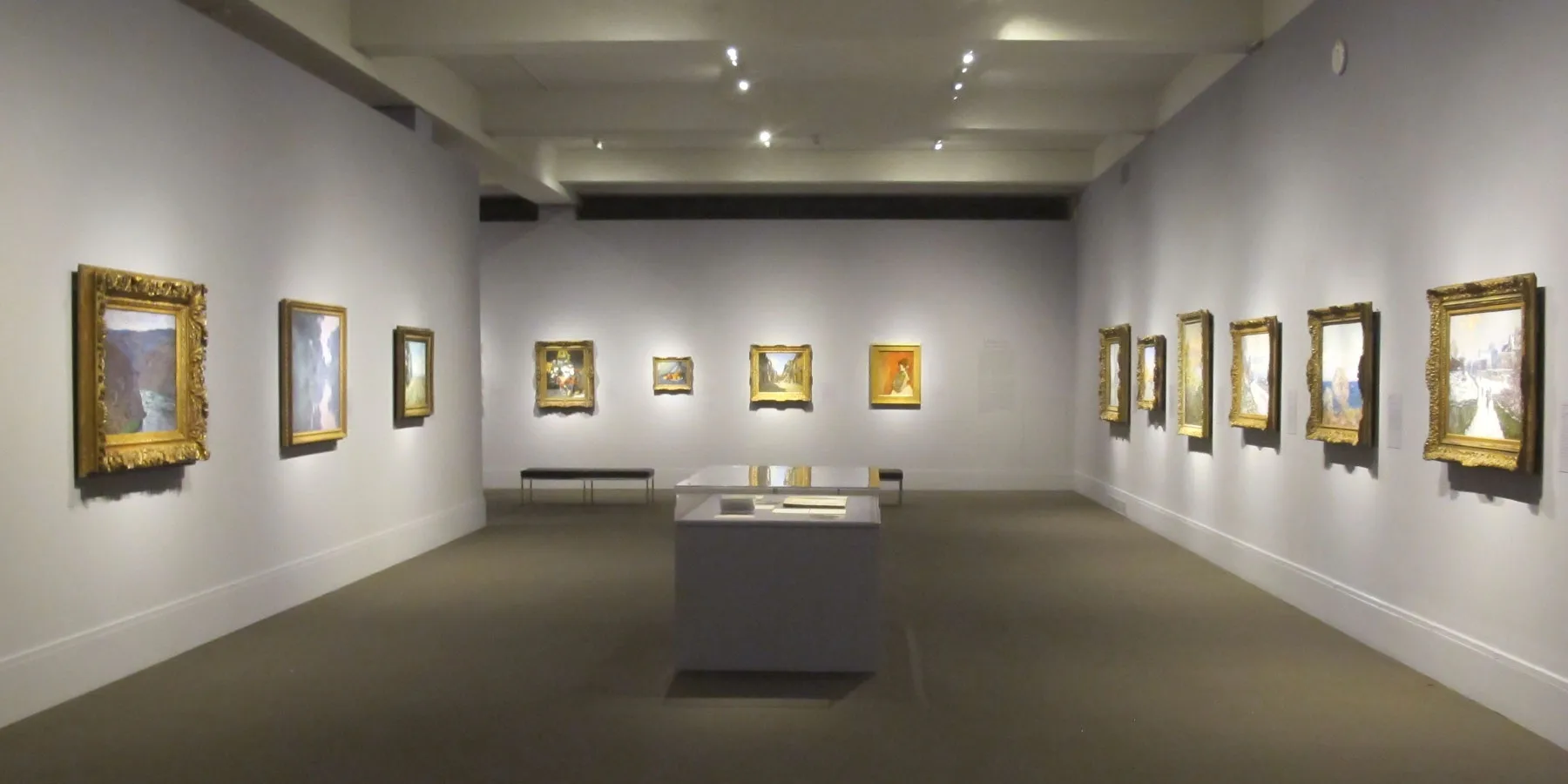 Minimalist gallery space with polished concrete floors, white walls, and recessed ceiling lights displaying framed artworks in ornate gold frames by Michiru Tanaka Lighting Design.