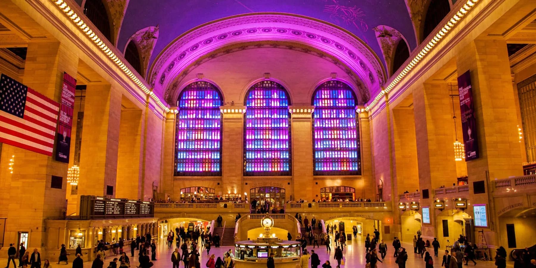 Grand Central Terminal's main concourse features magenta and purple LED lighting on the barrel vault ceiling and arched windows, with golden architectural elements and crowds of visitors below, designed by Michiru Tanaka Lighting Design.