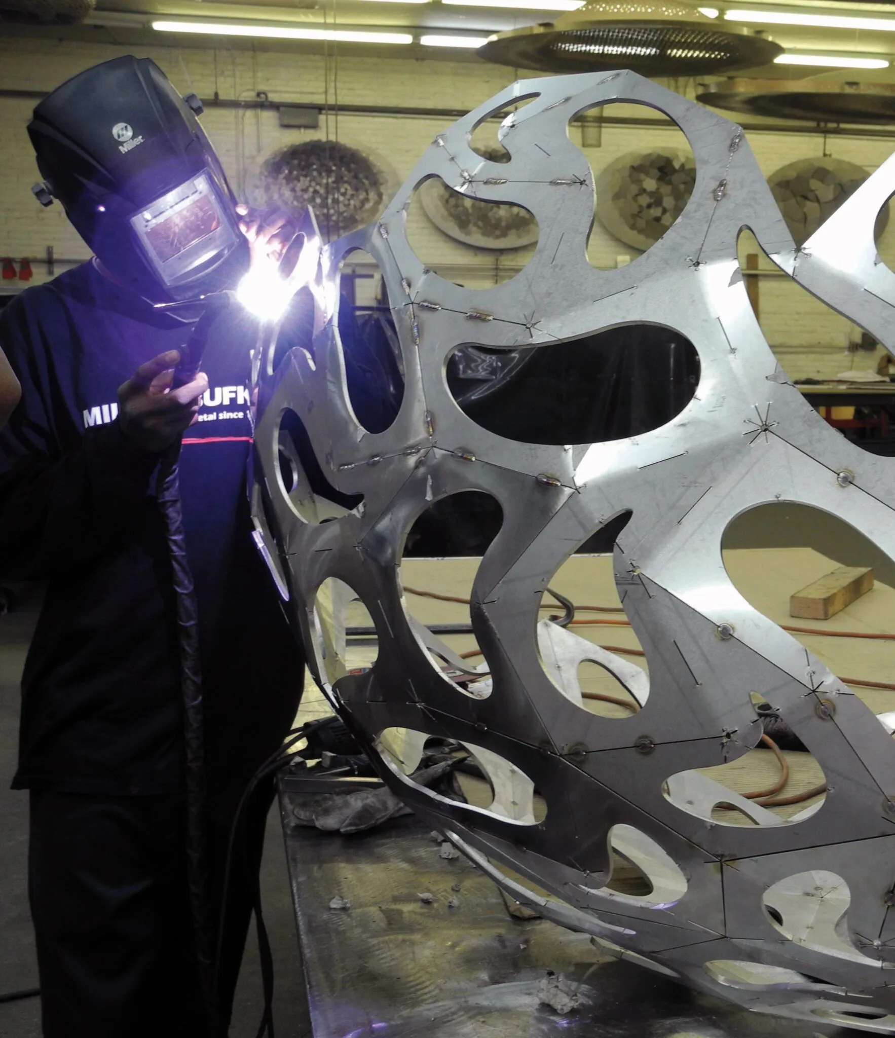 Welder in protective gear fabricating a large white perforated metal sculptural installation with organic curved forms in an industrial workshop by Milgo/Bufkin.