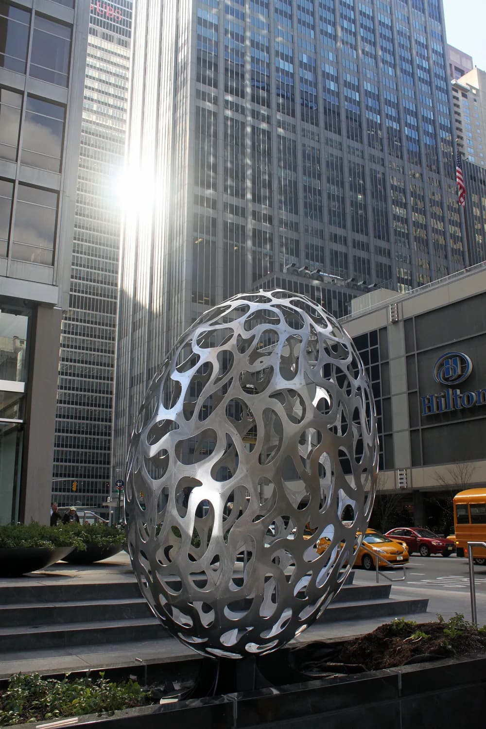 Large-scale perforated metal sphere sculpture installed in an urban plaza among skyscrapers, featuring organic oval cutouts and polished aluminum finish by Milgo/Bufkin.