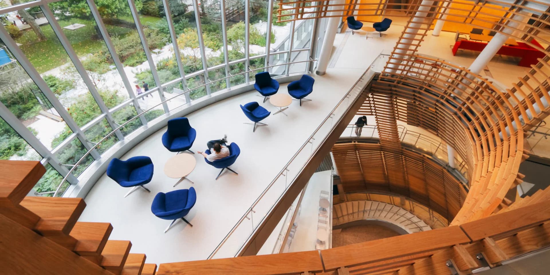 Modern multi-story atrium with blue chairs arranged on white platform, wooden curved stairs, and expansive glazed walls overlooking greenery, designed by Mitchell Giurgola.