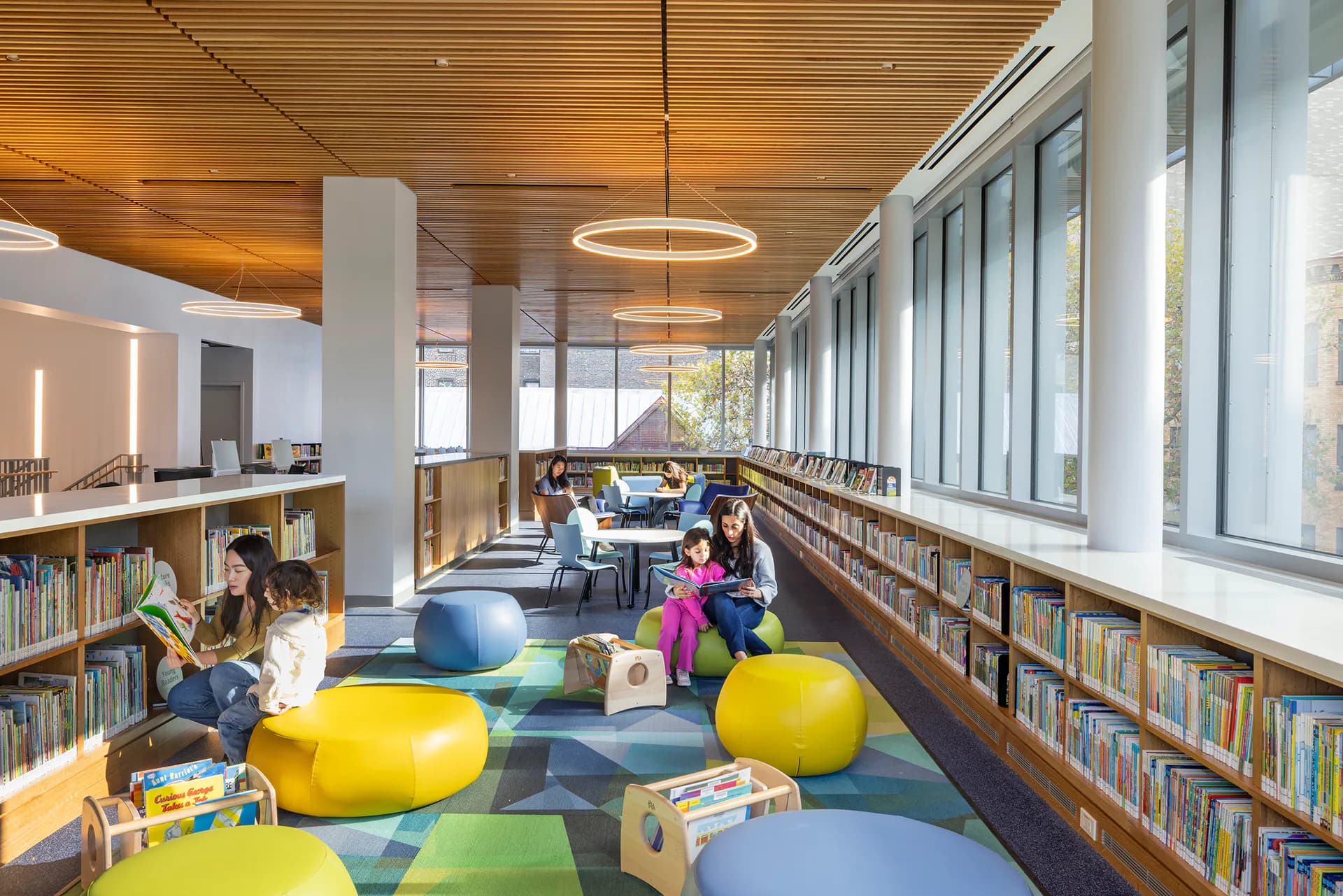 Modern library interior with colorful floor seating, built-in bookshelves, natural light, and suspended circular lighting, designed by Mitchell Giurgola.