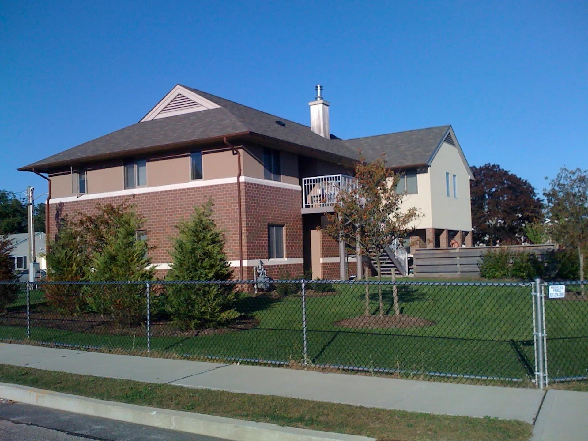 Residential home with red brick facade and white upper section, enclosed by chain-link fence, featuring modern pitched roof design by Mitropoulos Architects.