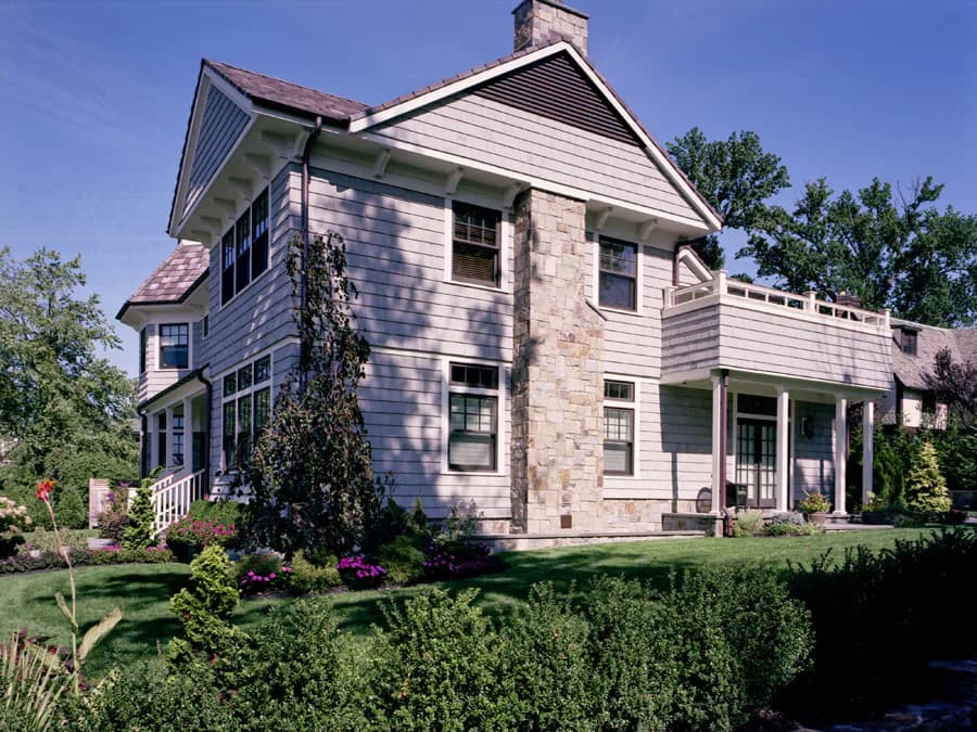 Two-story residential home with mixed stone and siding exterior, featuring covered porches, gabled roof, and manicured landscaping by Mitropoulos Architects.