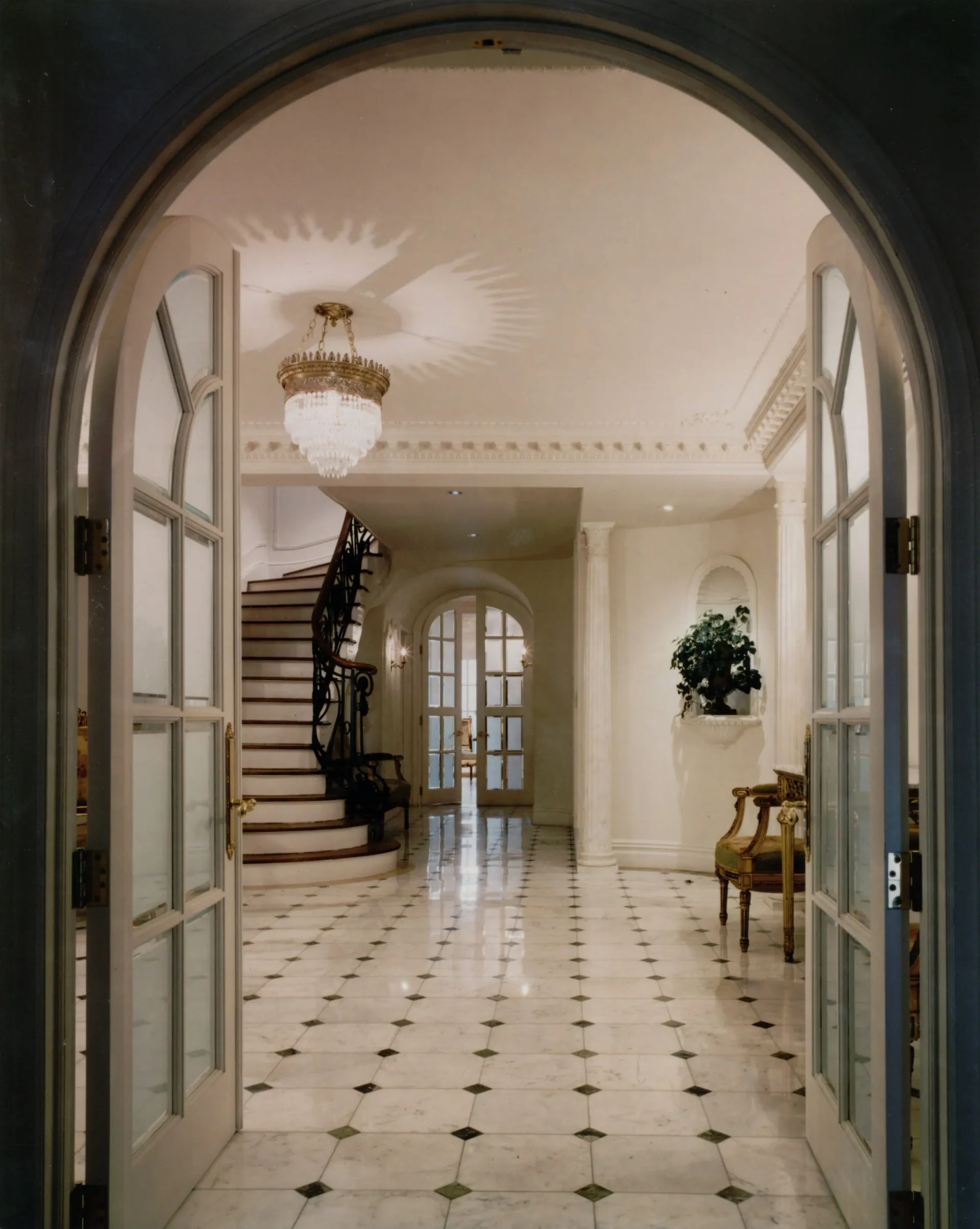 Elegant entrance hall with arched doorway, marble checkerboard flooring, brass chandelier, and curved staircase by MJM+A architects, PLLC.