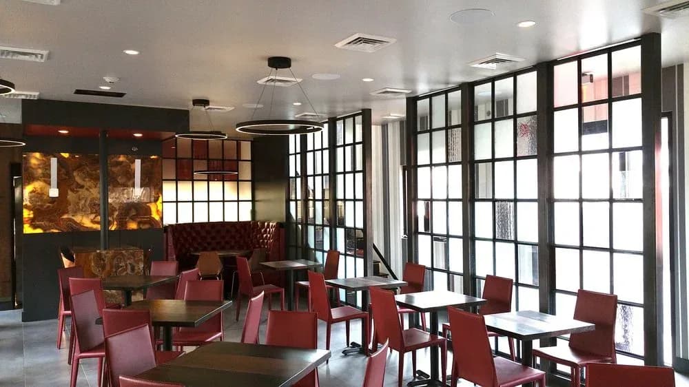 Modern restaurant or lounge interior with burgundy chairs, dark wood accents, geometric grid partition screens, warm ambient lighting, and floor-to-ceiling windows, designed by MJM+A architects, PLLC.