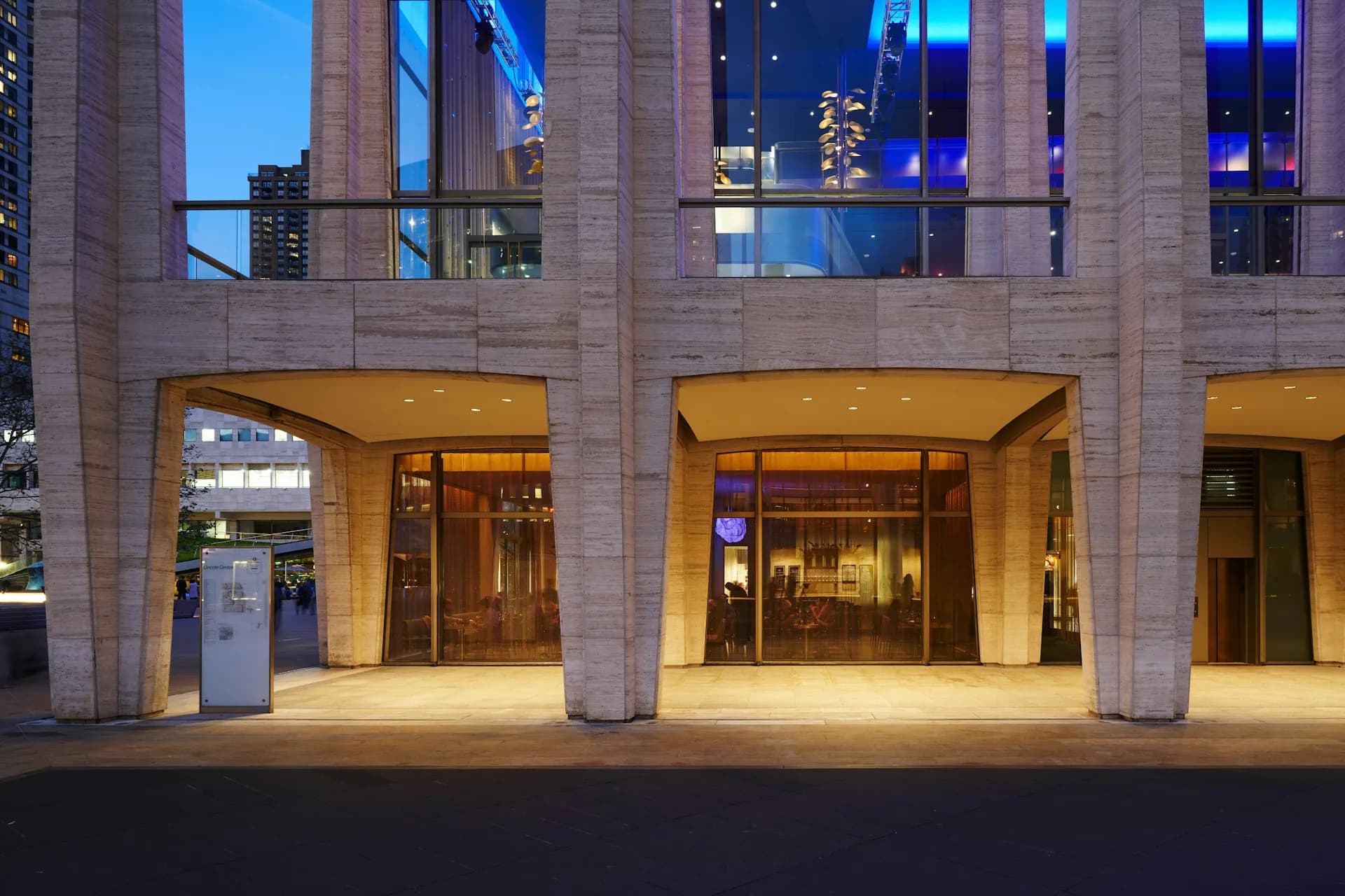 Illuminated ground-floor retail storefronts with warm interior lighting, concrete columns, and blue-tinted upper facade at dusk by Modellus Novus.