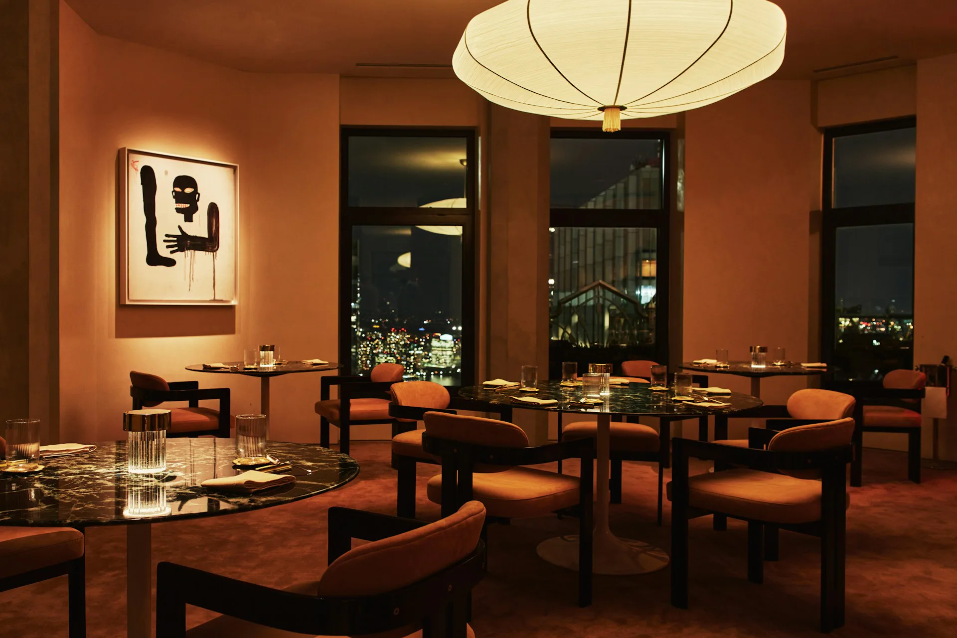 Warm orange-toned dining room with dark wood furniture, floor-to-ceiling windows overlooking city lights, geometric pendant lighting, and framed artwork by Modellus Novus.