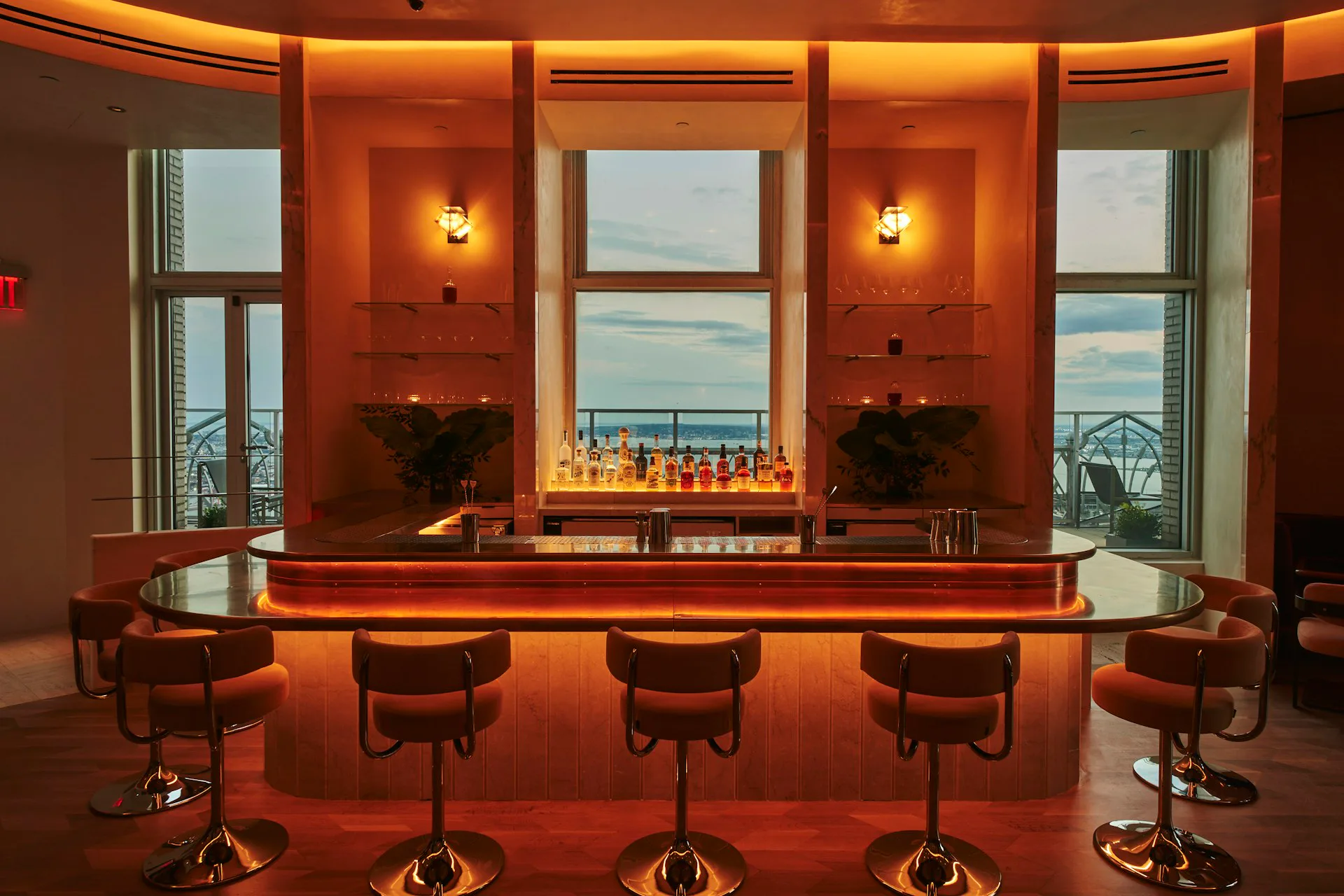 A luxurious bar interior features a curved amber-lit counter with five leather stools, warm orange walls, sconces, and expansive windows overlooking water by Modellus Novus.