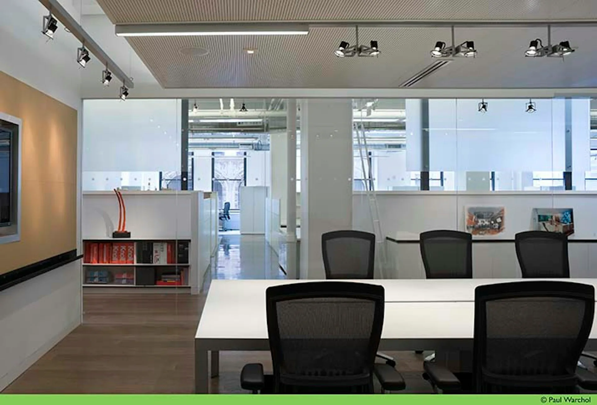 Modern office meeting room with white glass partitions, dark mesh chairs, white table, wood flooring, and track lighting, designed by Montroy Andersen DeMarco.