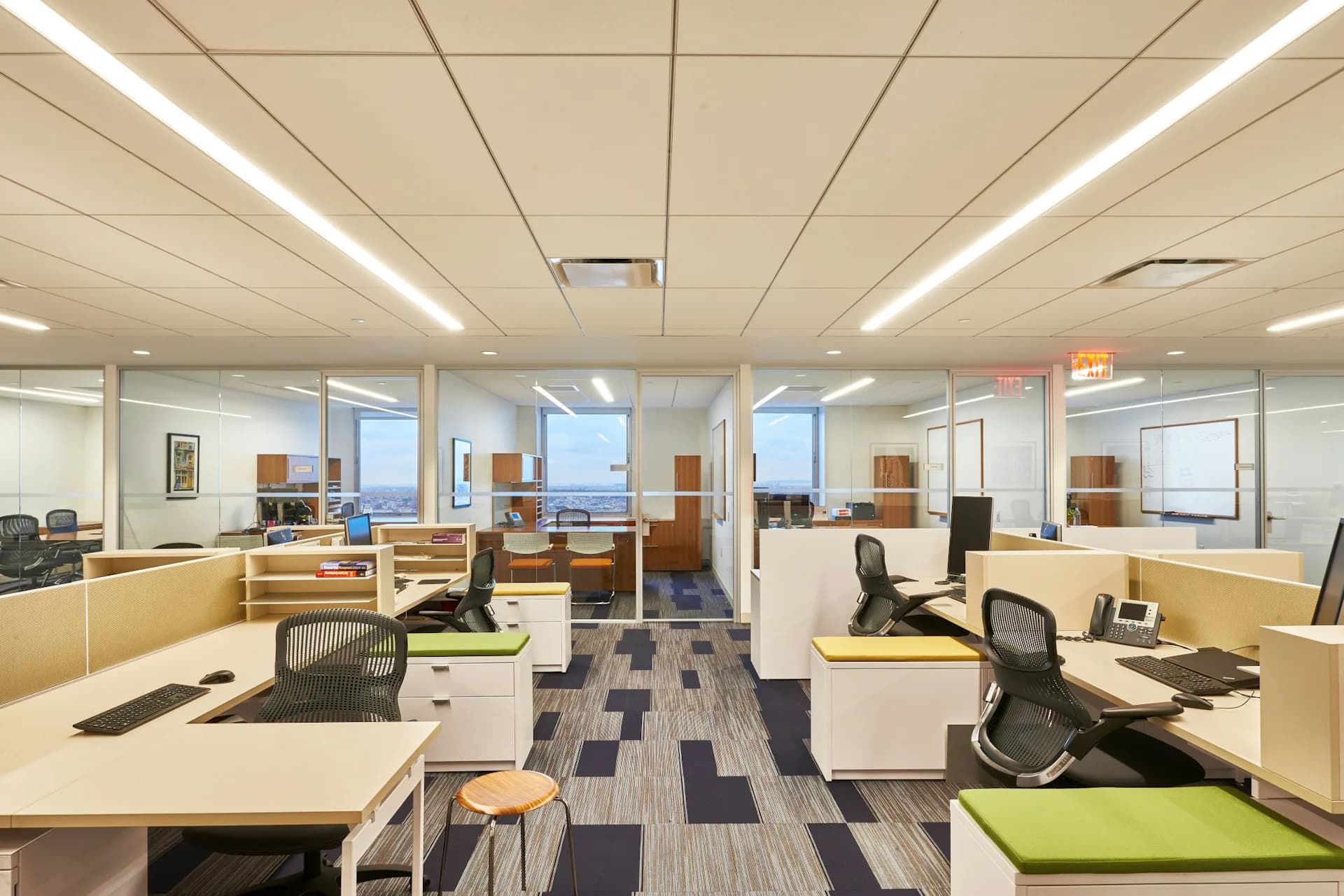 Modern open-plan office with natural wood desks, black mesh chairs, blue patterned flooring, and floor-to-ceiling windows overlooking a city skyline by Montroy Andersen DeMarco.