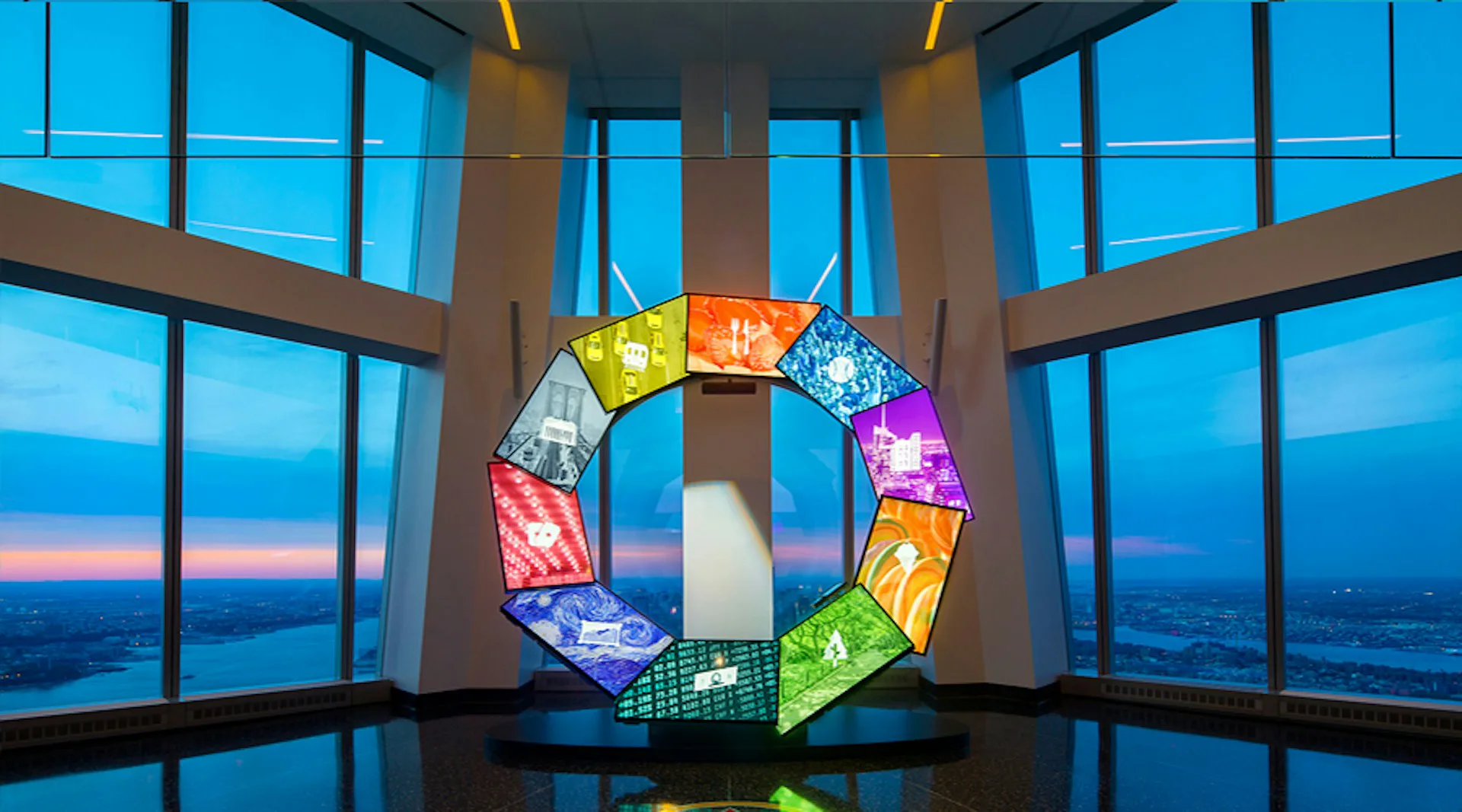 Modern circular installation with illuminated colorful panels suspended in a glass-enclosed high-rise space overlooking a city skyline at dusk, by Montroy Andersen DeMarco.