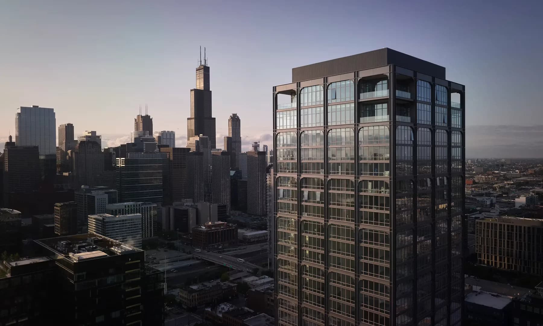 Chicago skyline at dusk showing a modern dark-glazed residential tower in the foreground with the illuminated downtown cityscape and Willis Tower visible in the background, designed by Morris Adjmi Architects.
