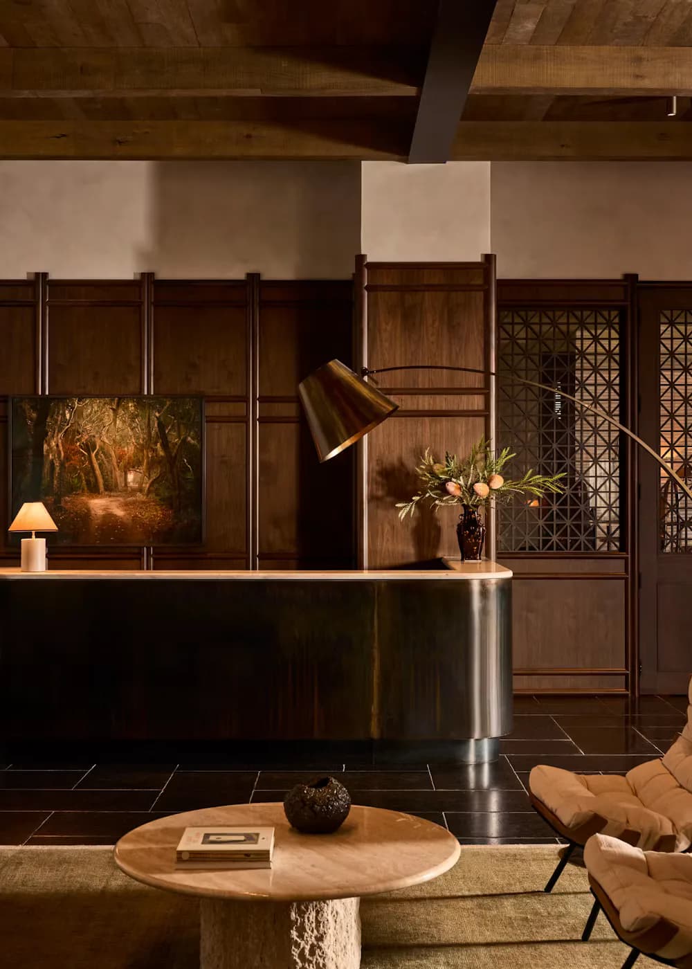 Contemporary hotel lobby with warm wood paneling, stainless steel reception desk, brass task lighting, and modernist furnishings in earthy tones by Morris Adjmi Architects.