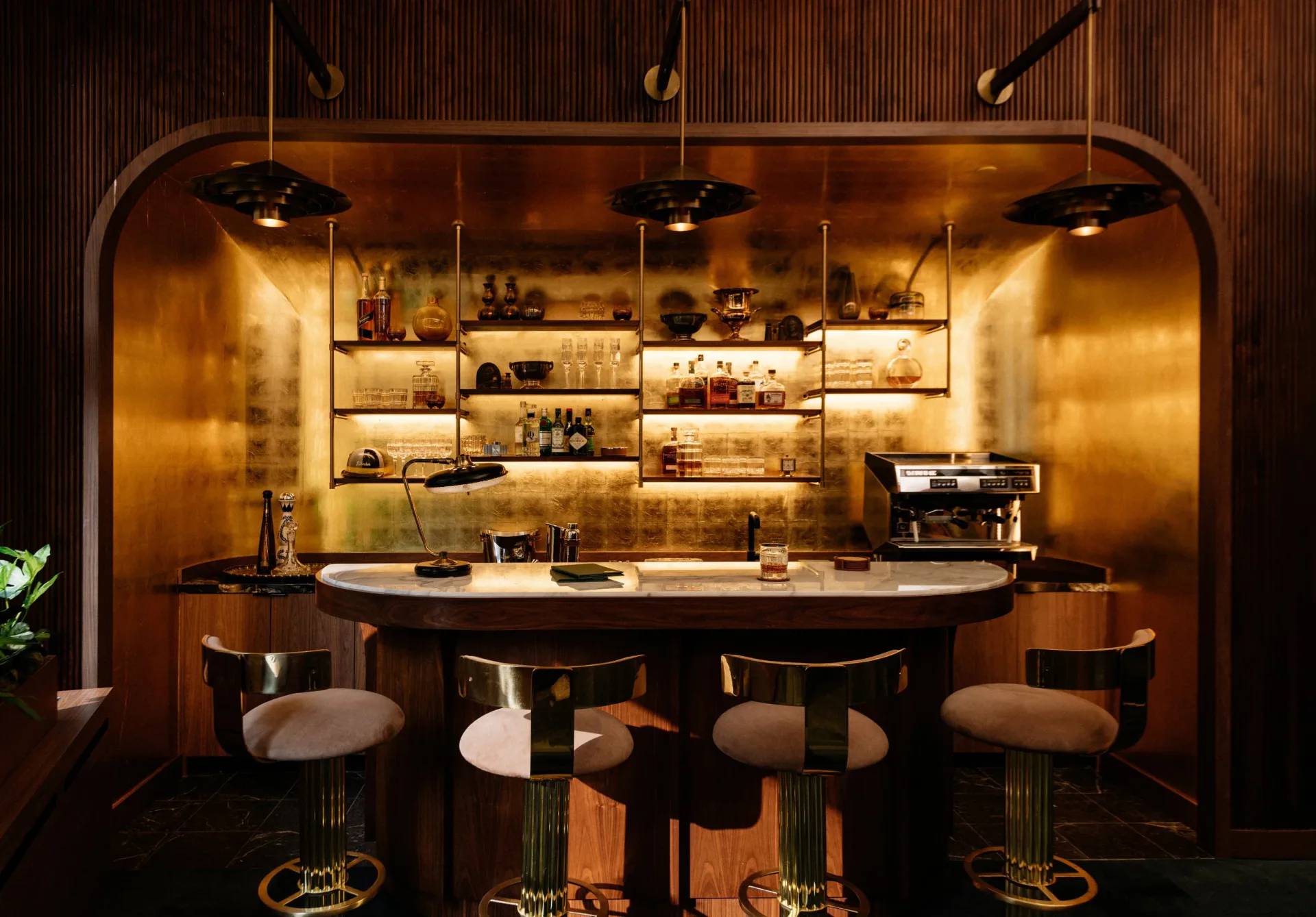 Warm-toned bar interior with brass-backed shelving, black curved counter, pendant lighting, and upholstered stools by MRD Lighting.