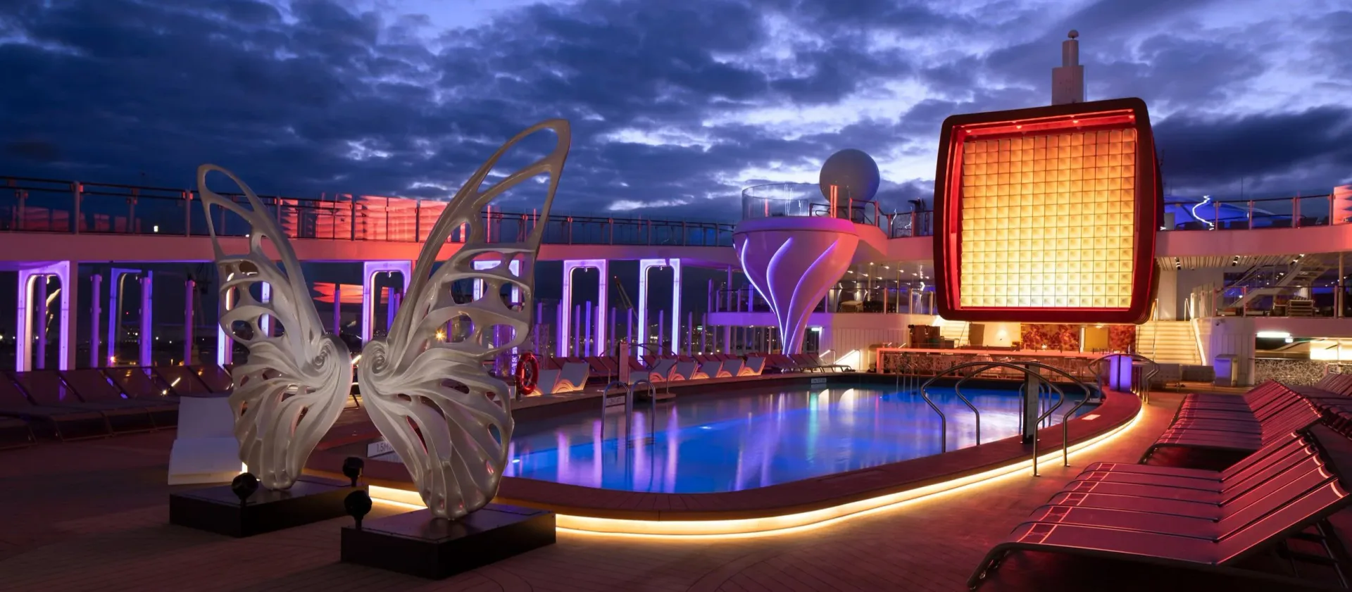 Illuminated waterfront entertainment complex with purple and orange neon lighting, sculptural elements, and reflective pool at dusk by MRD Lighting.