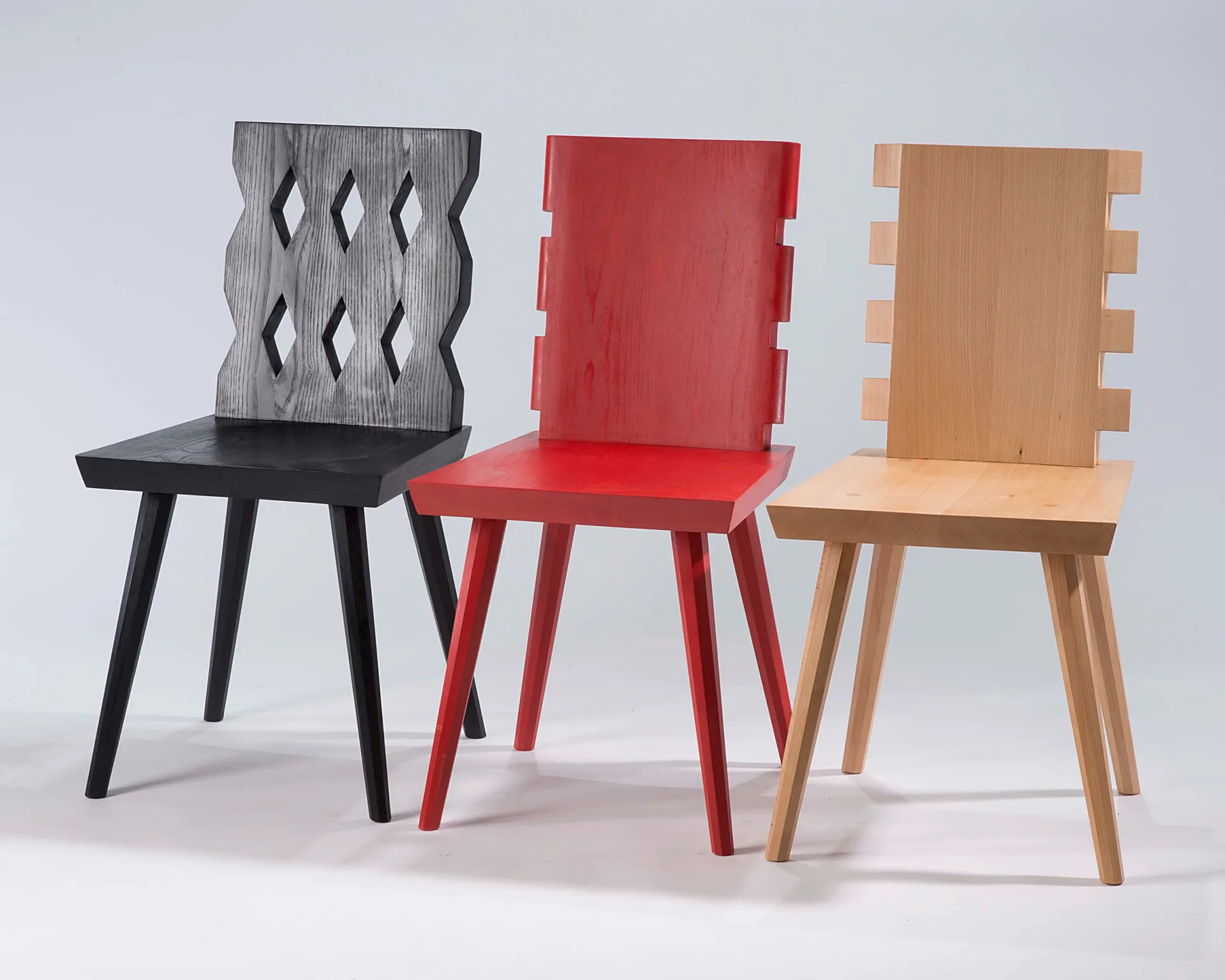 Three modernist chairs with geometric cutout backs in black, red, and natural wood finishes, arranged against a white background by MTM Studio.
