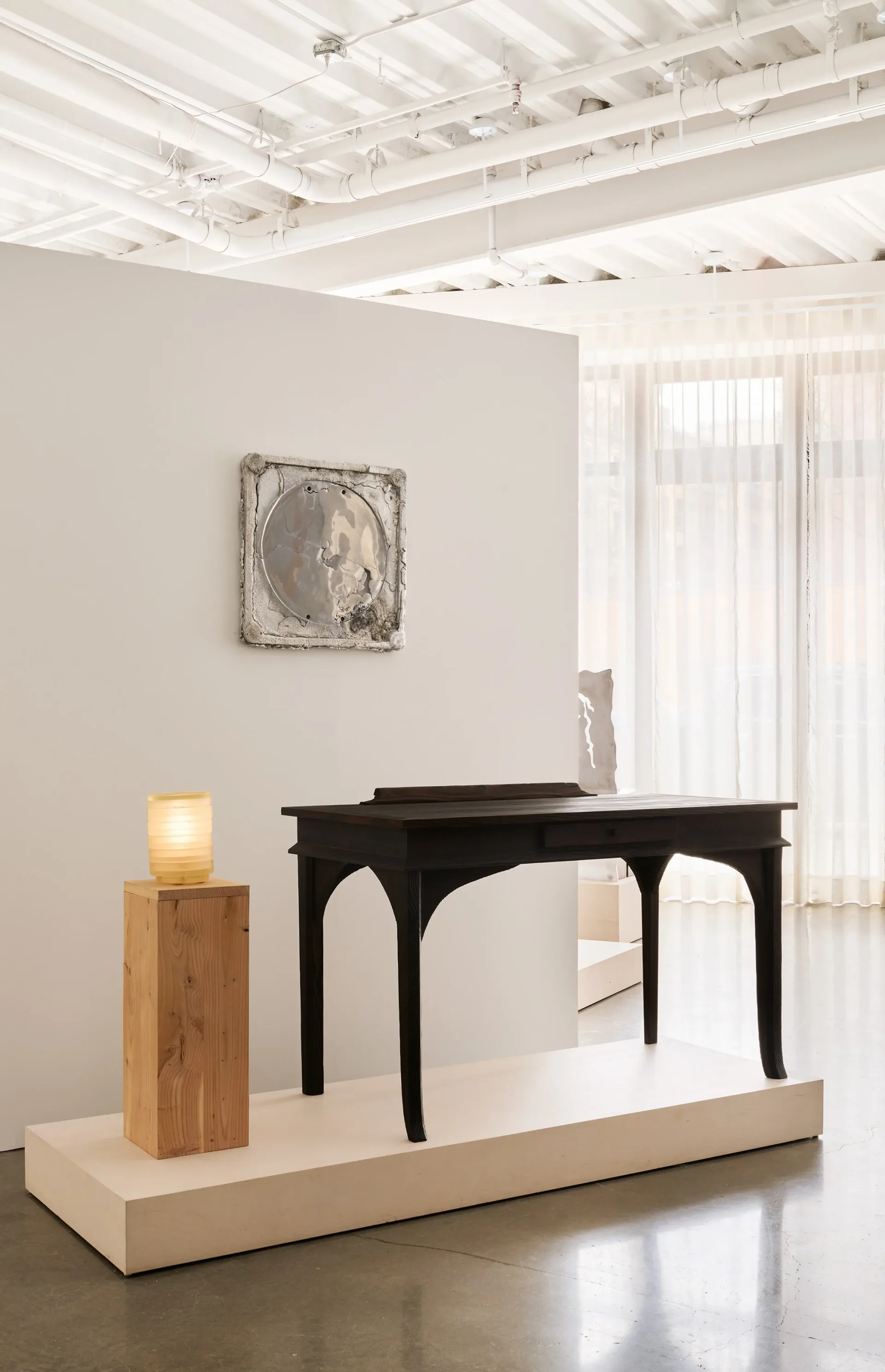 Minimalist exhibition space with white walls and exposed ceiling, featuring a black console table and wooden pedestal with warm lighting, designed by MTM Studio.