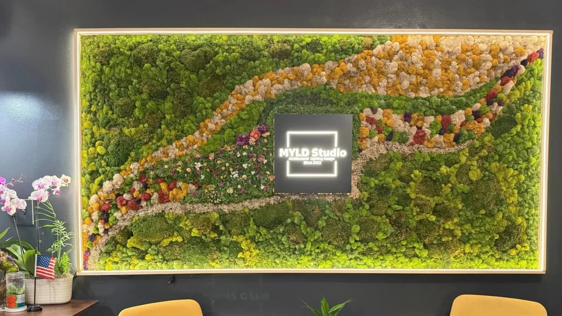 Living room wall installation featuring a large illuminated framed neon sign centered within an elaborate living moss and flower wall arrangement by MYLD Studio.