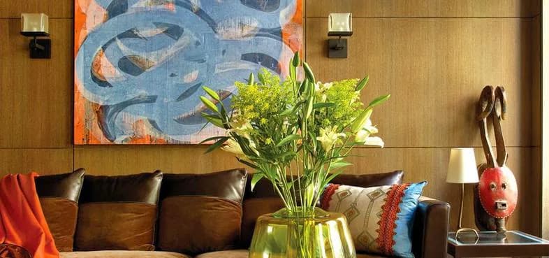 Modern living room with brown leather sofa, wooden paneling, blue abstract artwork, fresh floral arrangement, and colorful accent pillows by MZ Woodwork.
