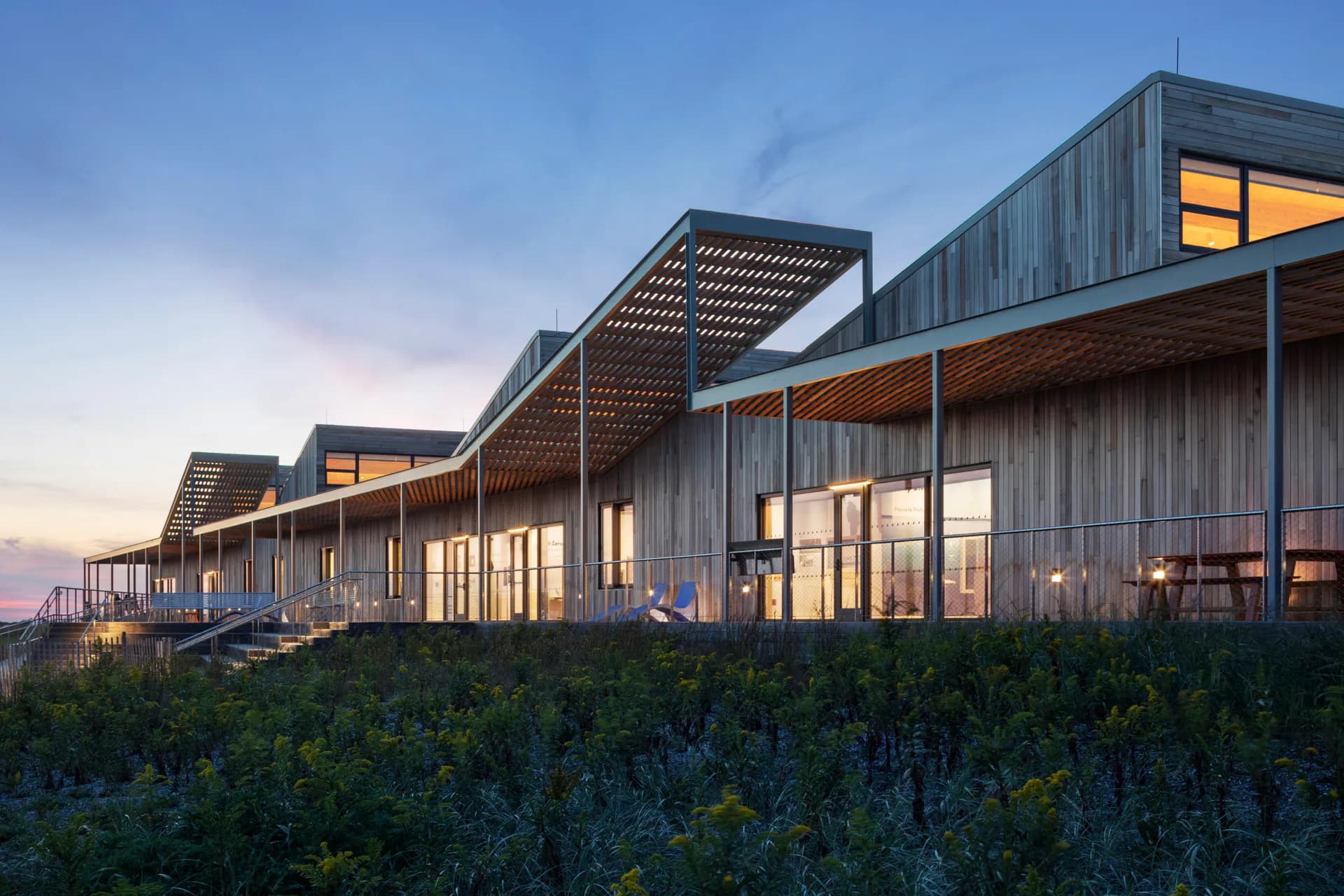 Modern residential buildings with dark wood cladding, expansive glazing, and pergola rooflines set against a twilight sky, surrounded by native vegetation, by nARCHITECTS.