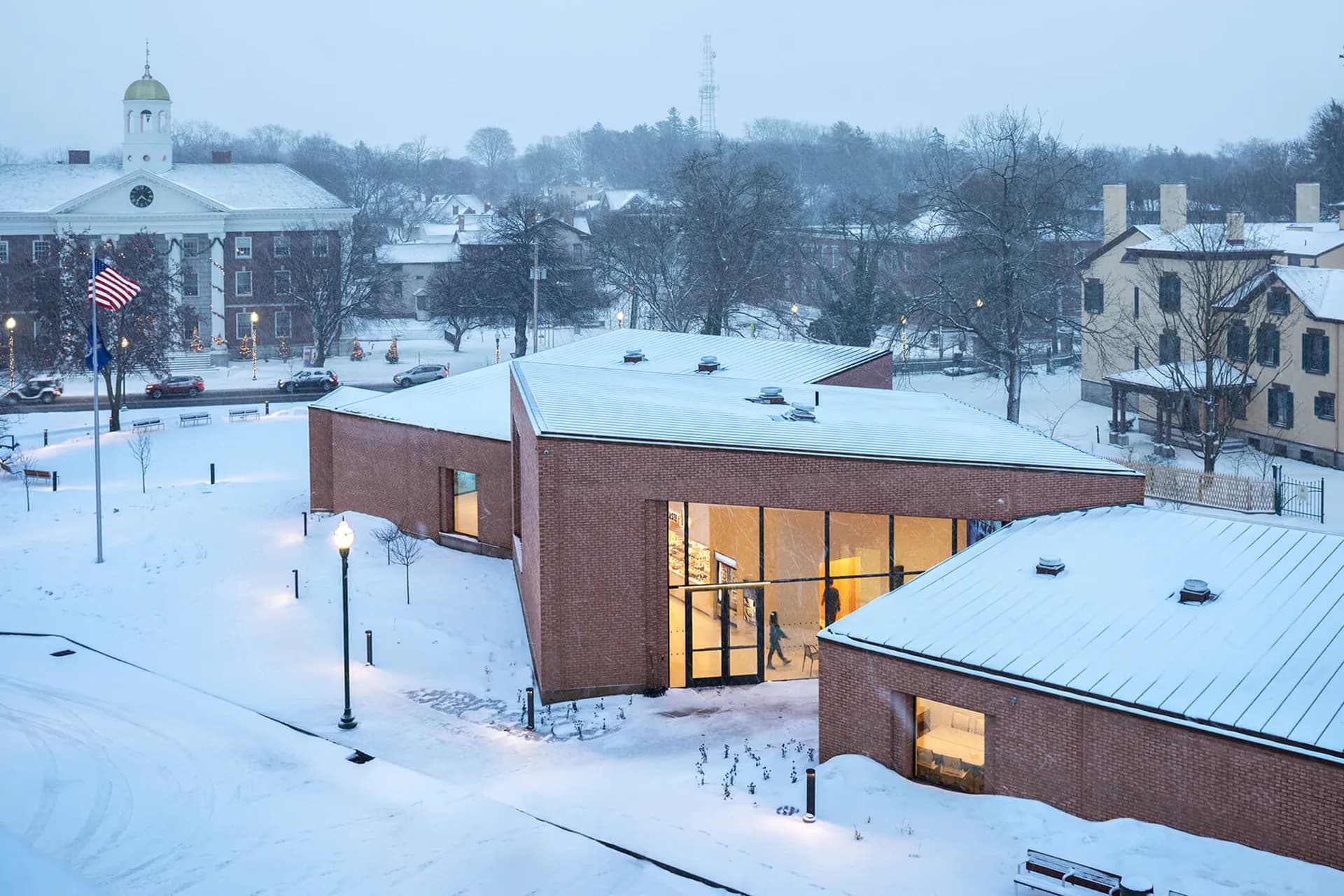 Modern brick pavilion with illuminated windows set in snowy campus landscape, featuring clean geometric forms and metal roofing by nARCHITECTS.