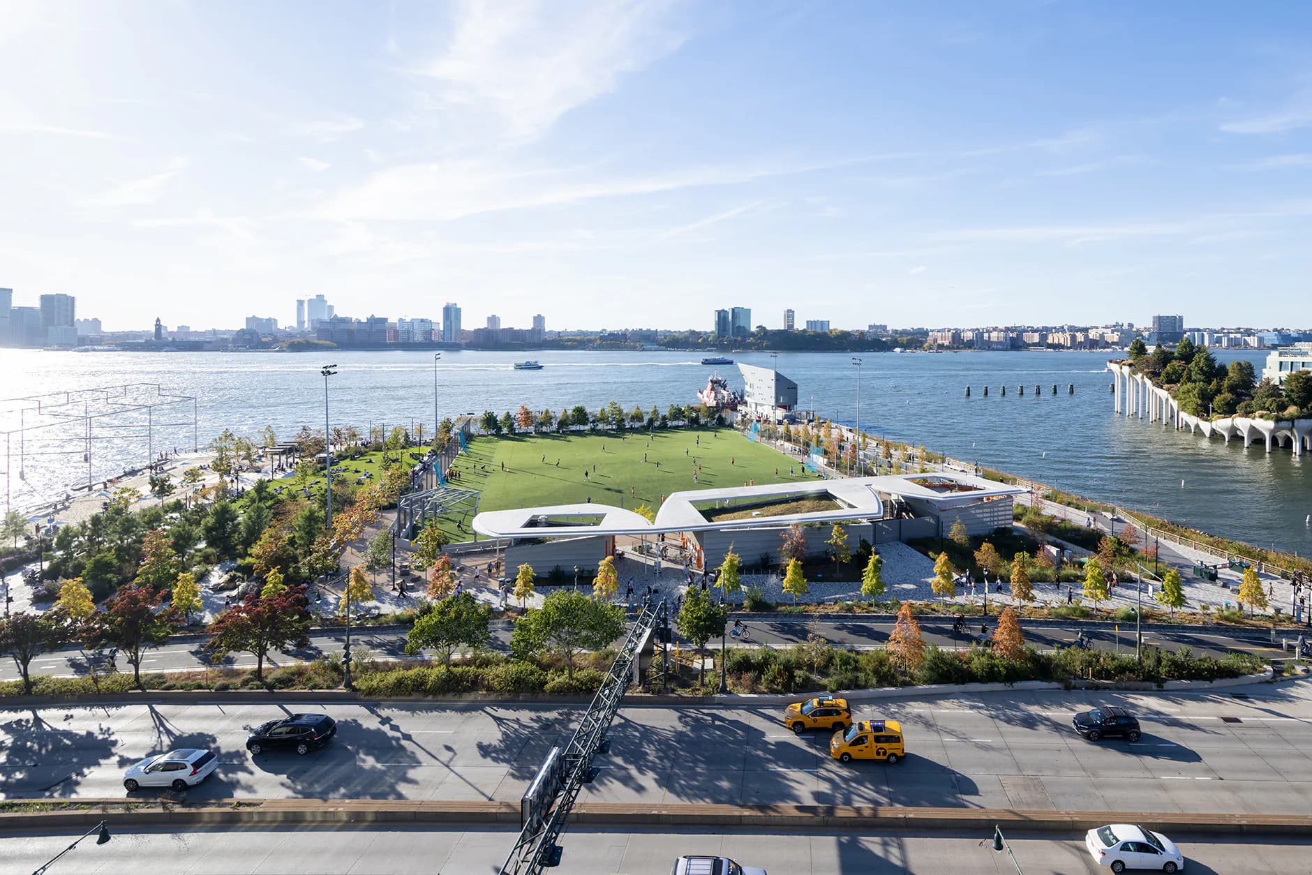 Waterfront public park with curved landscaping, green spaces, and modern waterside pavilion overlooking harbor with city skyline by nARCHITECTS.