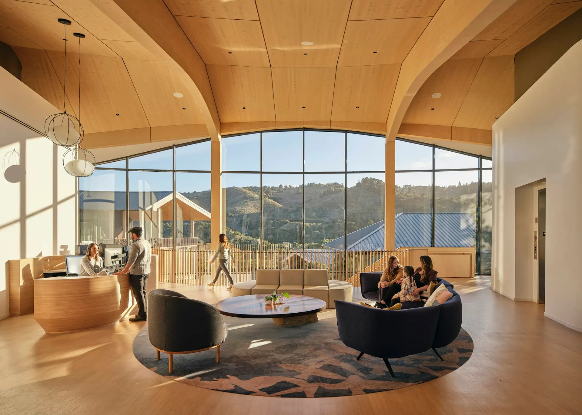 Modern institutional interior with curved wood ceiling, floor-to-ceiling windows overlooking mountains, black curved seating, and synthetic figures by NBBJ.