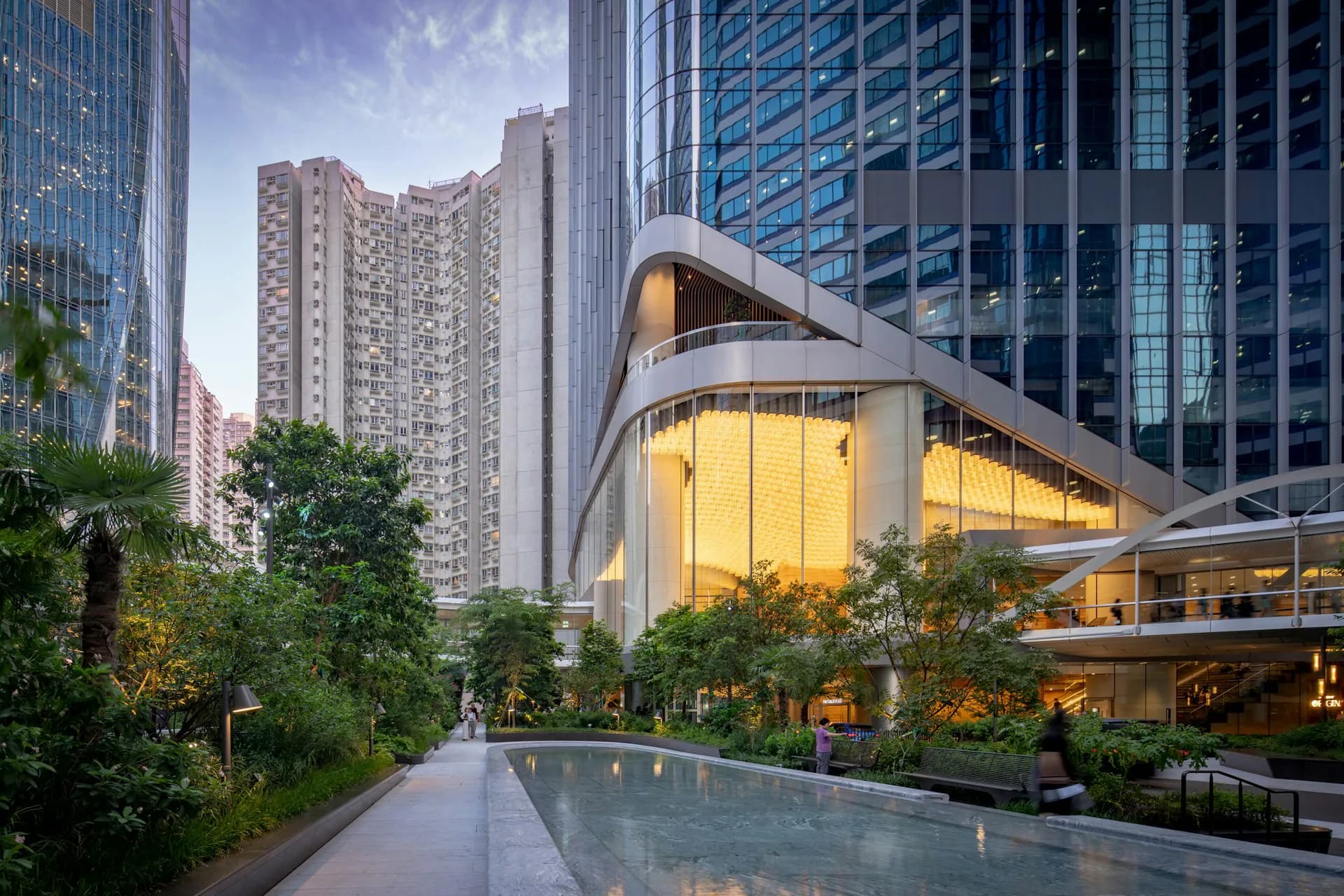 Modern curved building with illuminated yellow glass facade nestled among residential towers, featuring landscaped plaza with water feature by NBBJ.