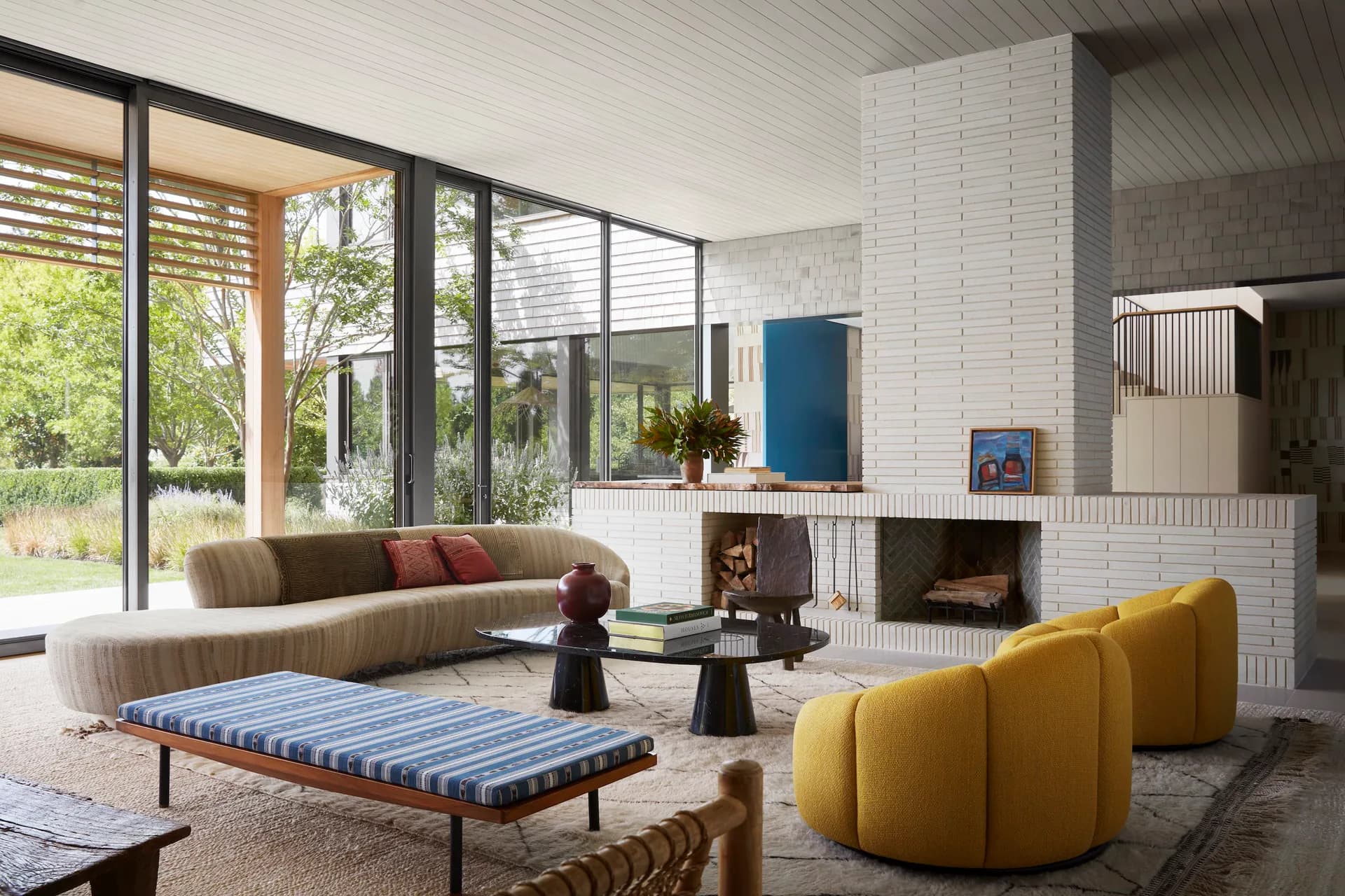 Modern living room with curved yellow seating, striped bench, white brick fireplace, and expansive glazing overlooking green landscape by Neal Beckstedt Studio.