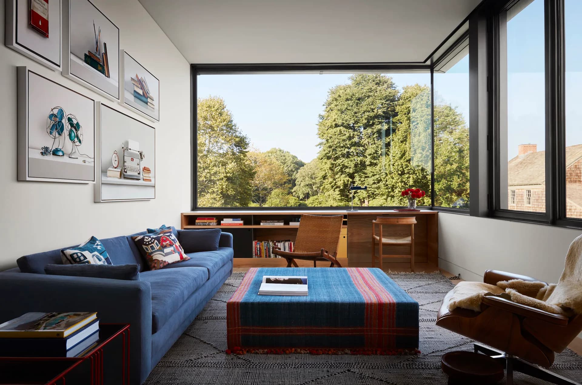 Modern living room with blue sofa, wood storage, striped ottoman, and floor-to-ceiling windows overlooking trees, designed by Neal Beckstedt Studio.