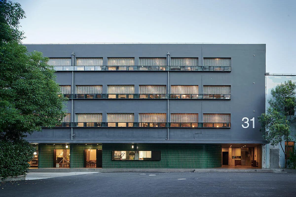 Modern residential building facade with stacked timber-screened balconies, dark green ground-floor storefront, and illuminated windows at dusk by Neri&Hu Design and Research Office.