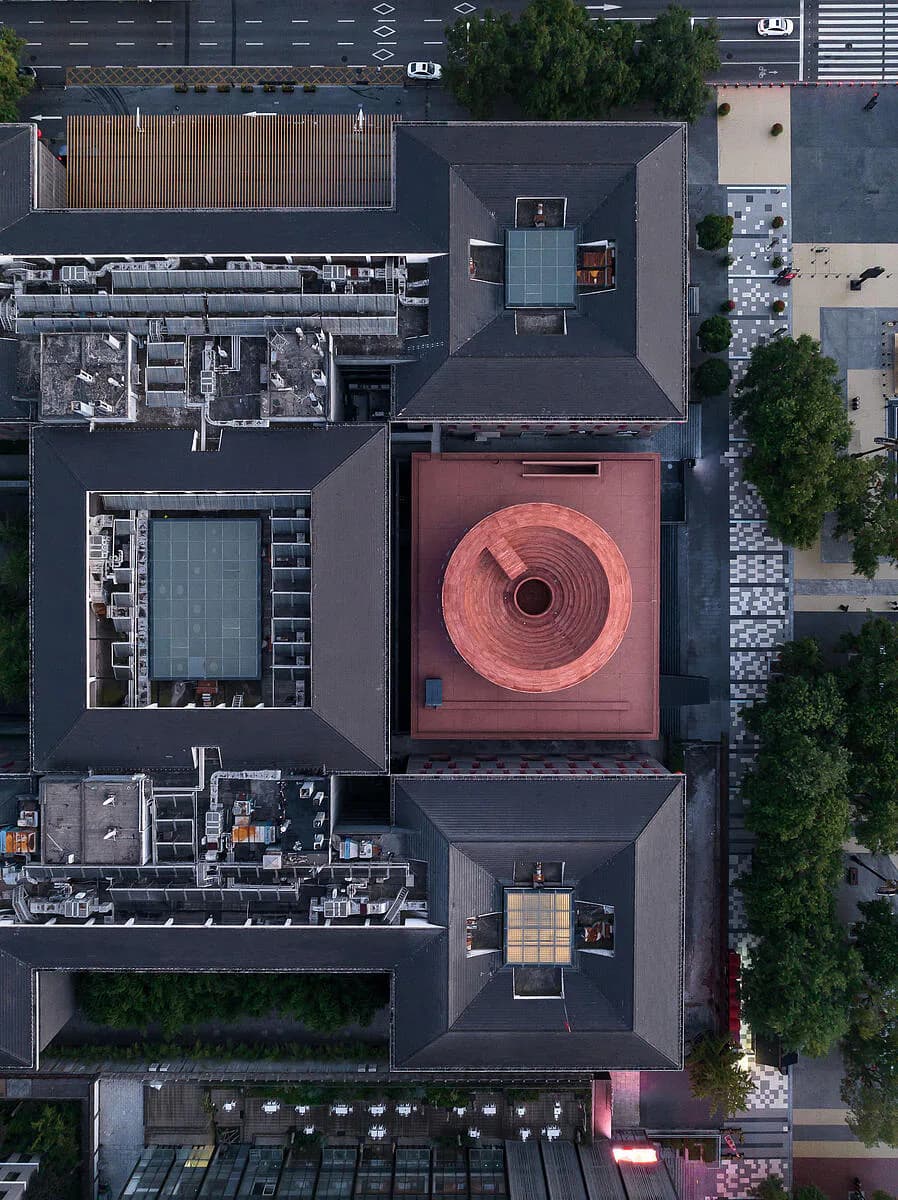 Aerial view of a contemporary courtyard design featuring a striking circular red installation, dark paving, and landscaped green spaces by Neri&Hu Design and Research Office.
