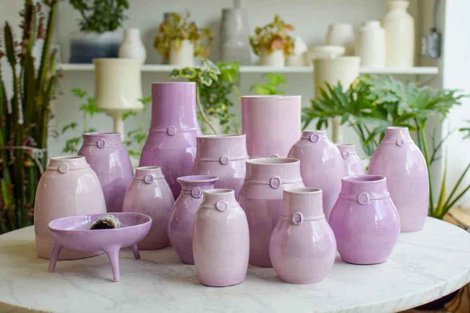 Collection of handcrafted ceramic vessels in soft mauve and pink tones arranged on a white surface, featuring various organic forms with subtle metallic accents, displayed against a backdrop of potted plants by Newcomb Studios (Nicholas Newcomb Pottery & Sculpture).