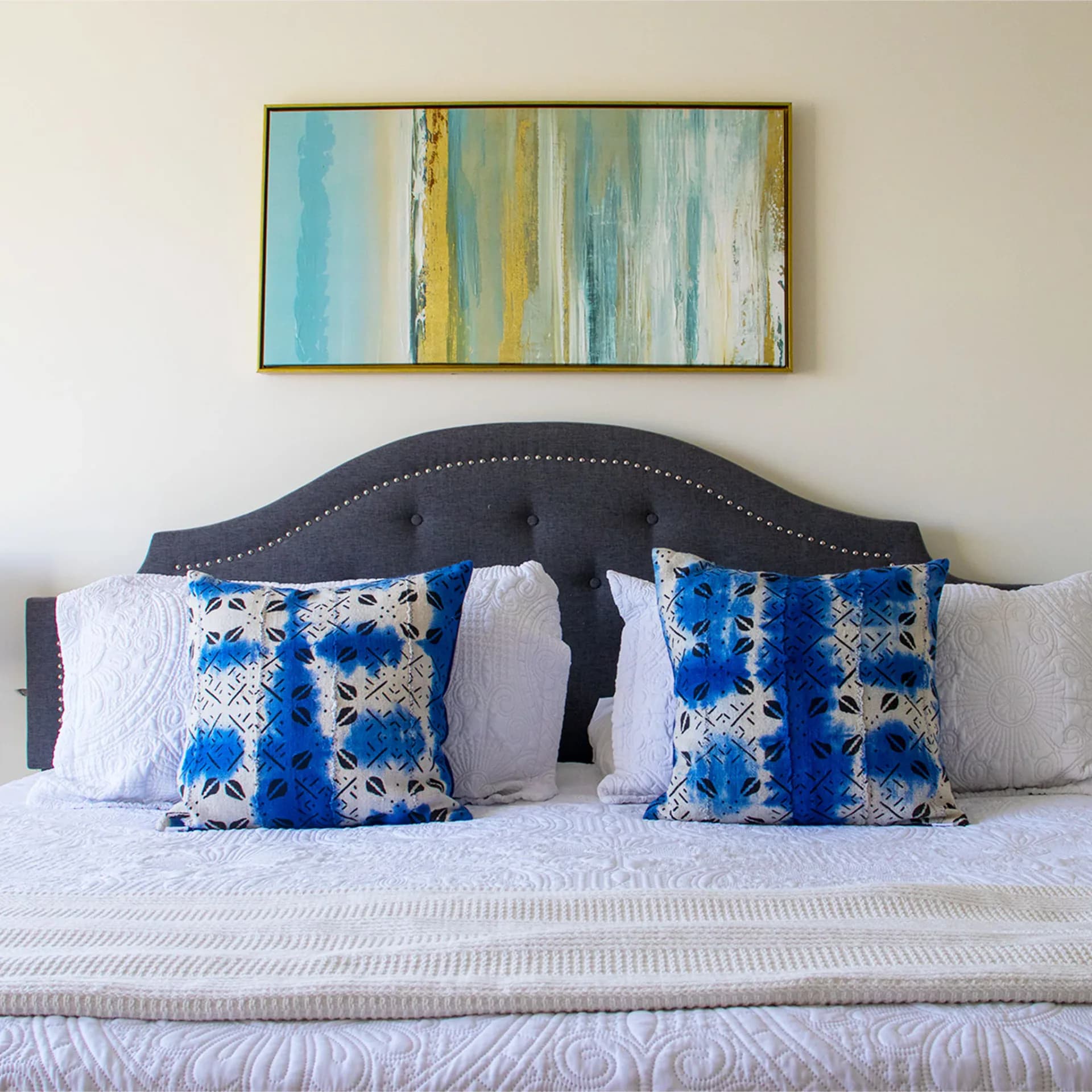 Bedroom featuring a dark upholstered headboard with nailhead trim, layered with blue patterned and white pillows, above a neutral bedframe by Niche Handmade Furniture.