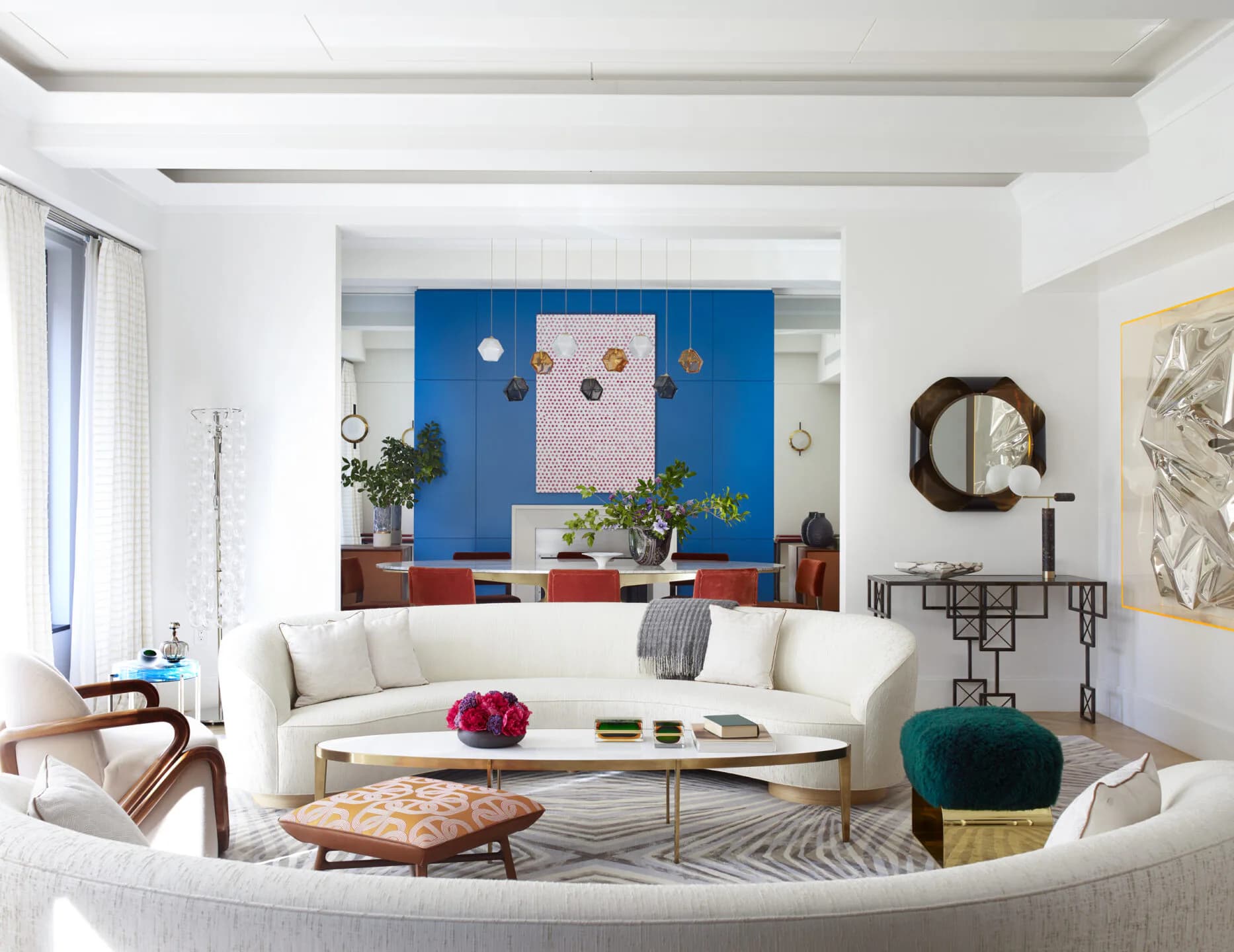 Modern living room with curved white sofa, blue accent wall with artwork, wooden side tables, patterned rug, and round mirror by Nicole Fuller Interiors.