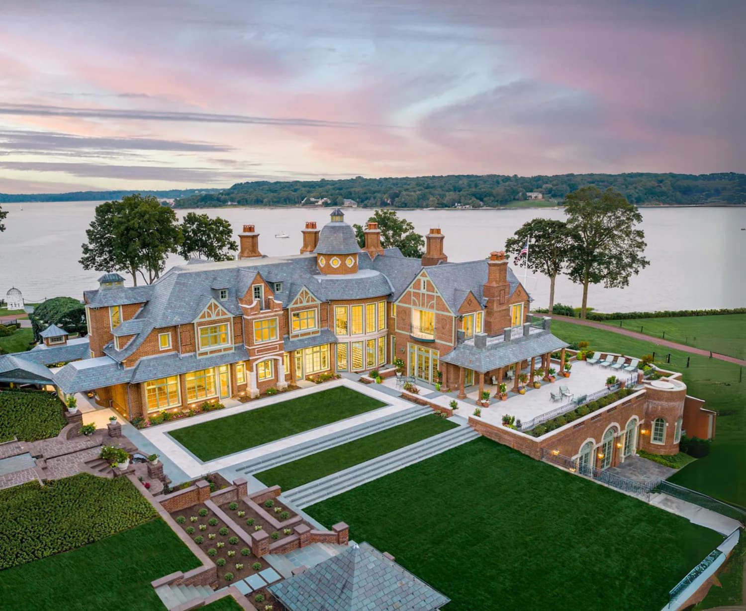 Waterfront luxury mansion with golden-yellow brick facade, slate roof, and manicured grounds featuring a modern pool pavilion, designed by NJ Caine Architecture.