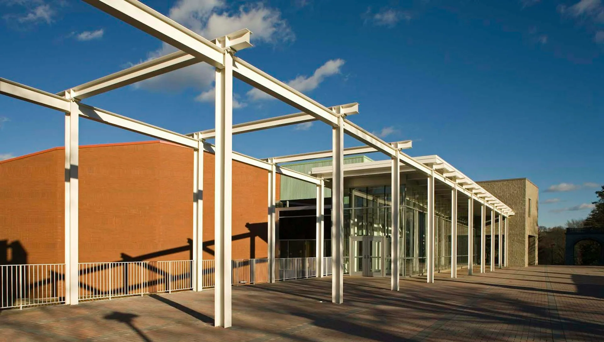 Modern institutional building with orange brick facade and white steel pergola structure, clear blue sky, minimal landscaping by NK Architects.